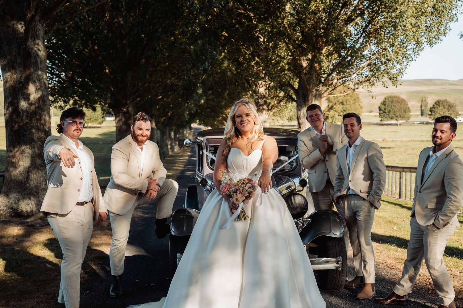 Bride in white gown posing with groom and groomsmen in beige suits beside a vintage car outdoors.