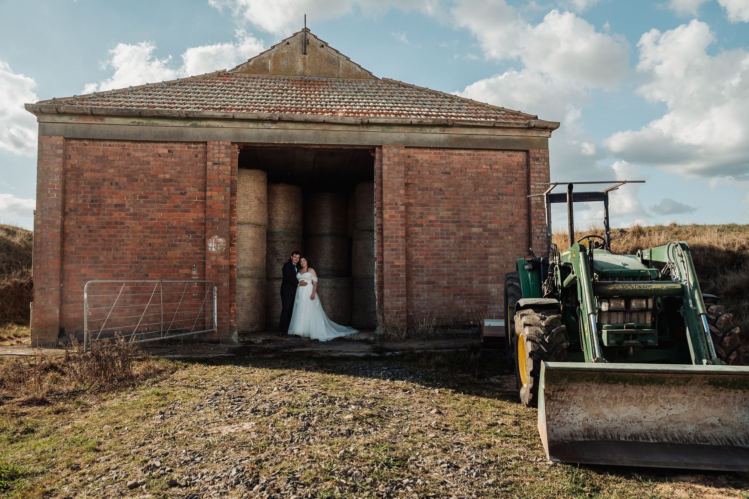 Bride and groom stand in the entrance of a rustic brick barn beside a vintage green tractor.