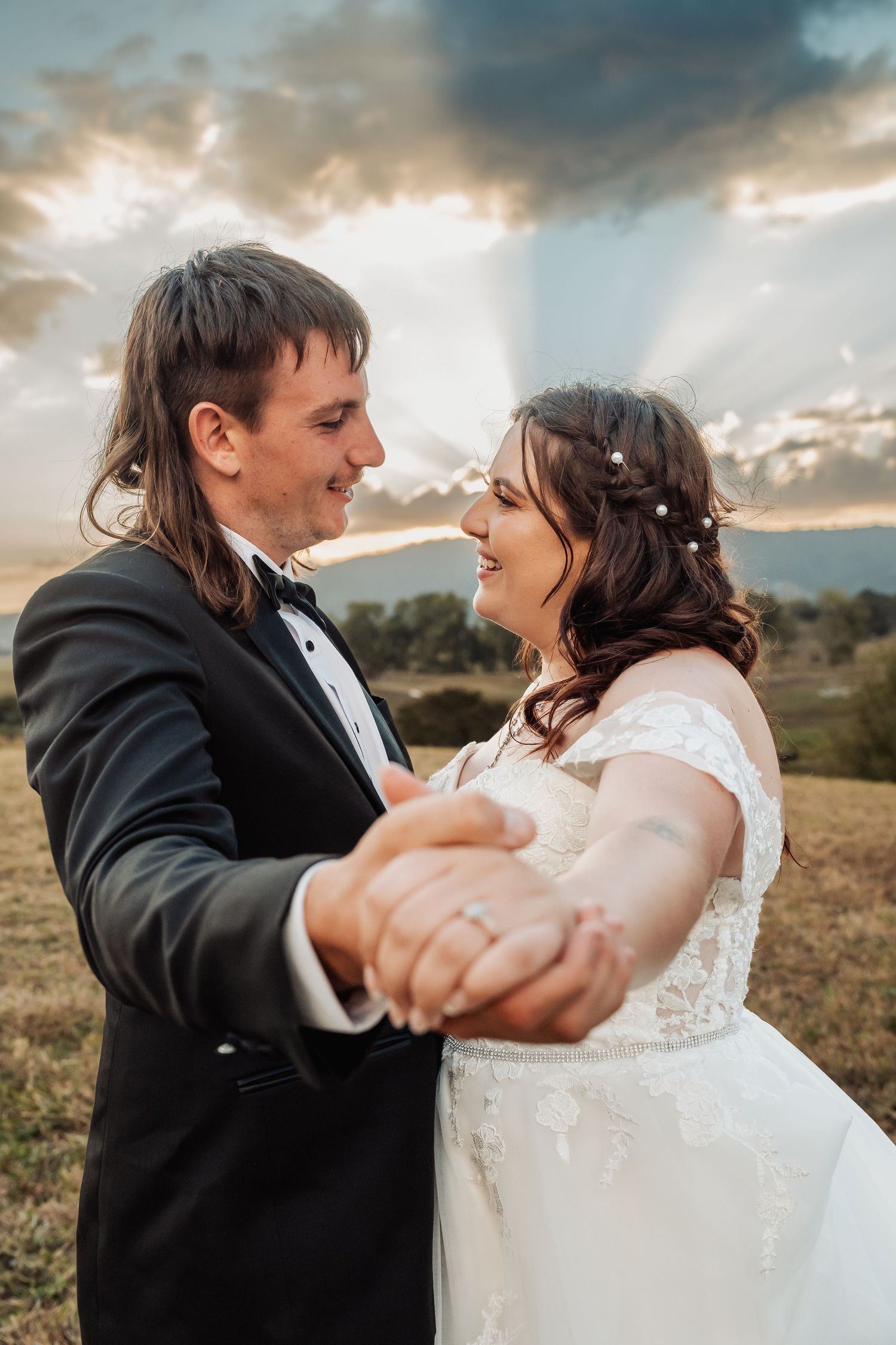 Smiling bride and groom holding hands dancing in golden field with dramatic sunset rays behind.
