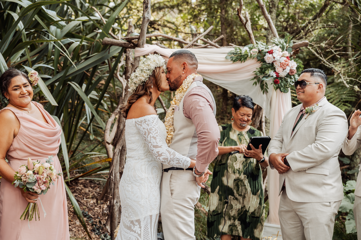 A wedding ceremony takes place under a floral arch with tropical greenery.