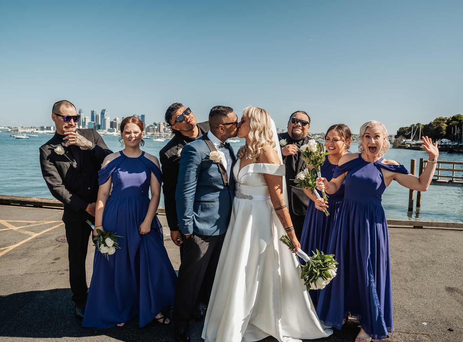 Wedding party poses together on pier with water and city views.