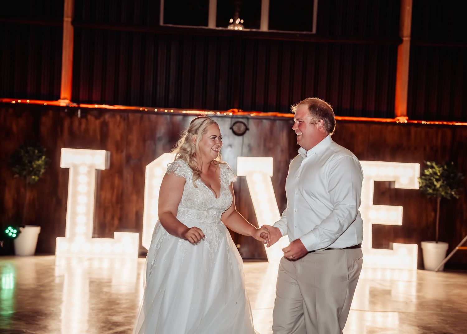 Bride and groom hold hands during first dance in front of illuminated LOVE marquee letters in rustic barn.