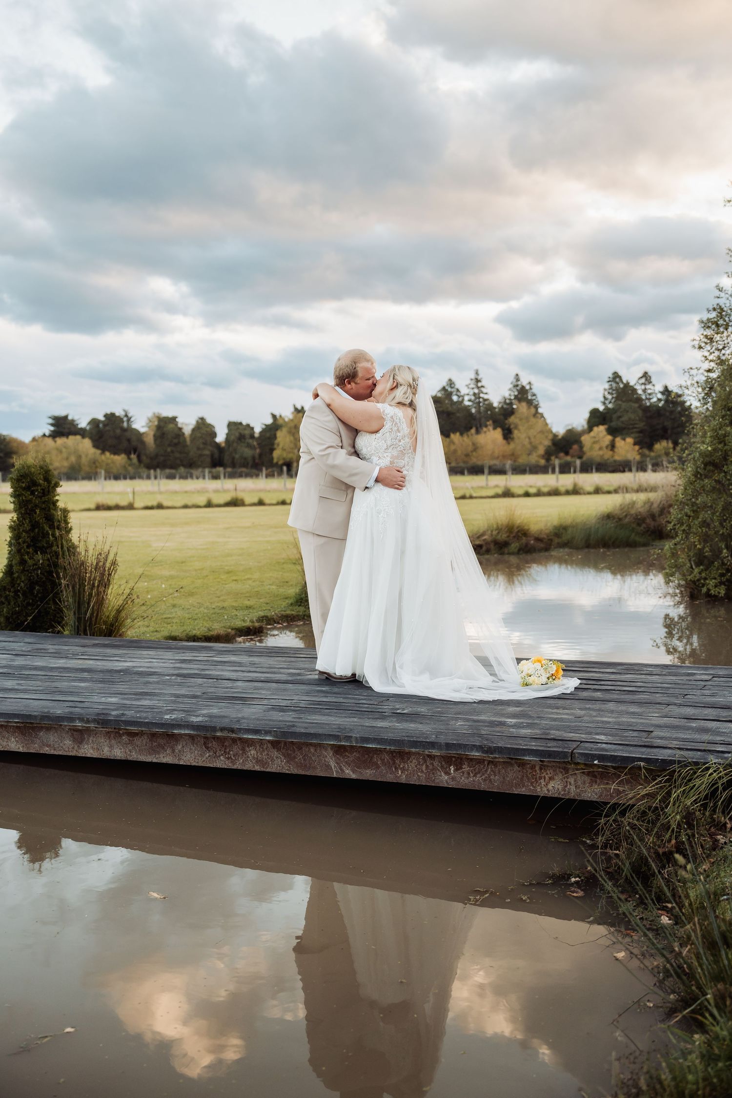 Bride and groom share romantic kiss on wooden bridge over pond reflecting cloudy sky.
