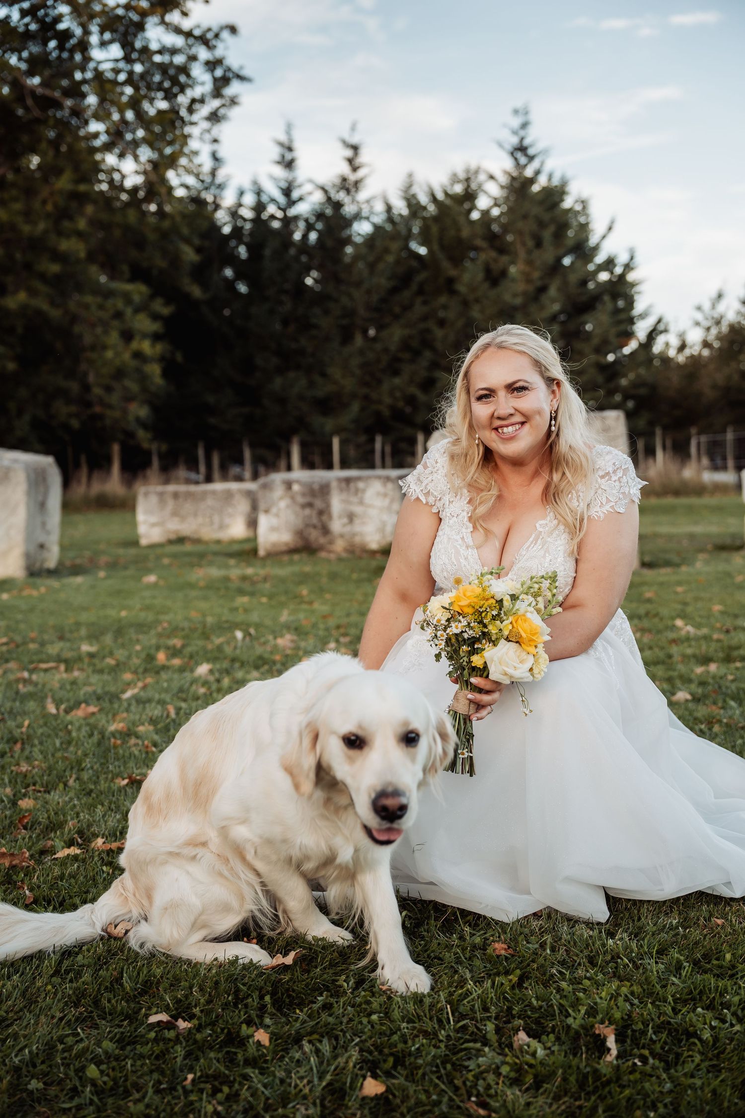 Smiling bride in lace wedding dress posing with a golden retriever dog while holding yellow bouquet.