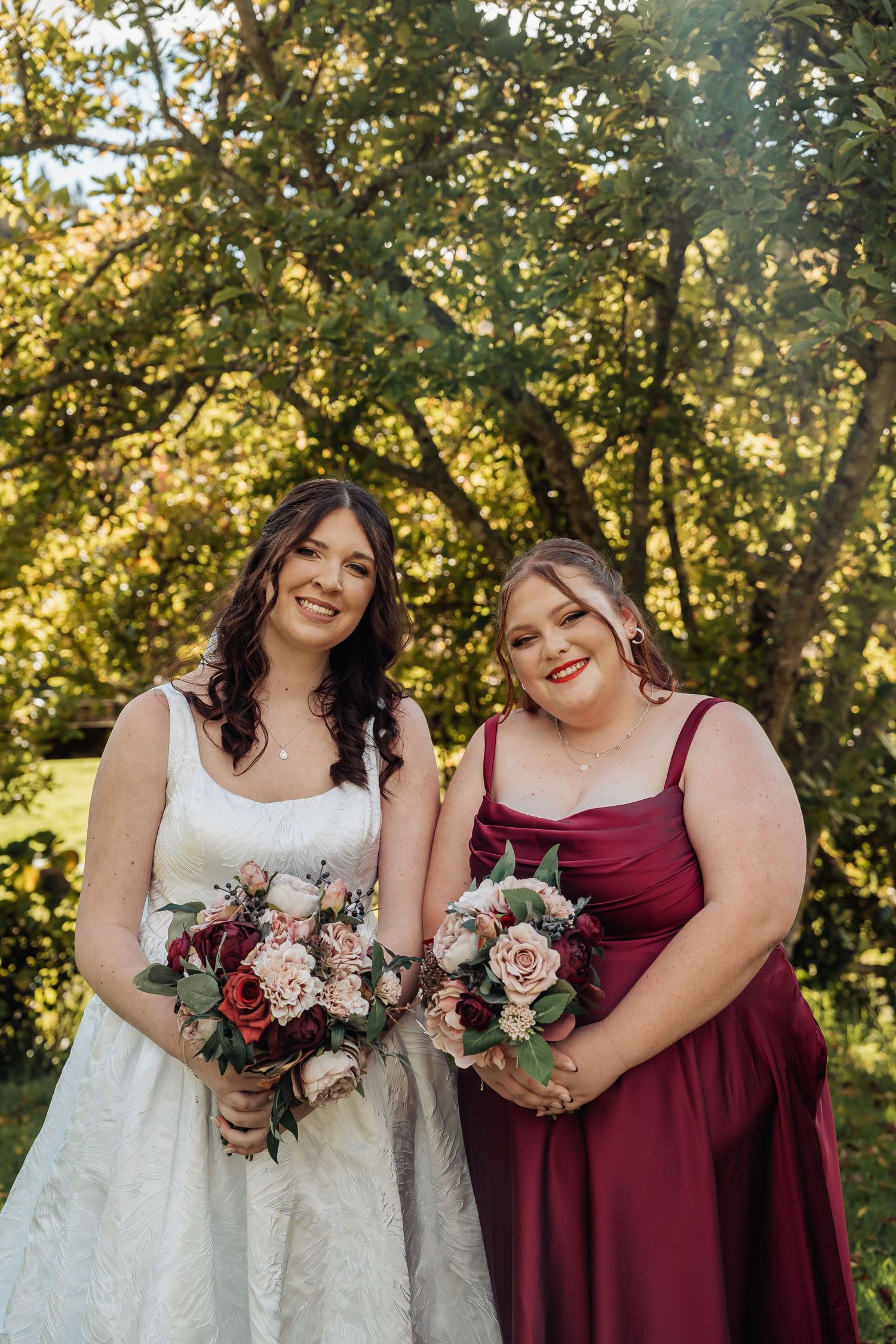 Bride in white lace gown and bridesmaid in burgundy dress holding floral bouquets outdoors.