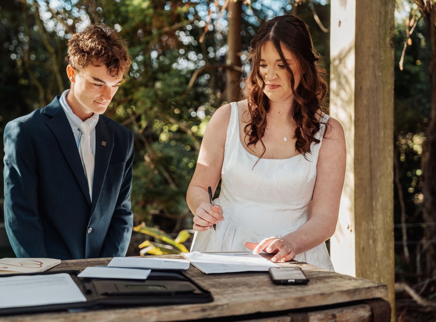 Bride signing wedding documents at rustic wooden table with groom in navy suit beside her.