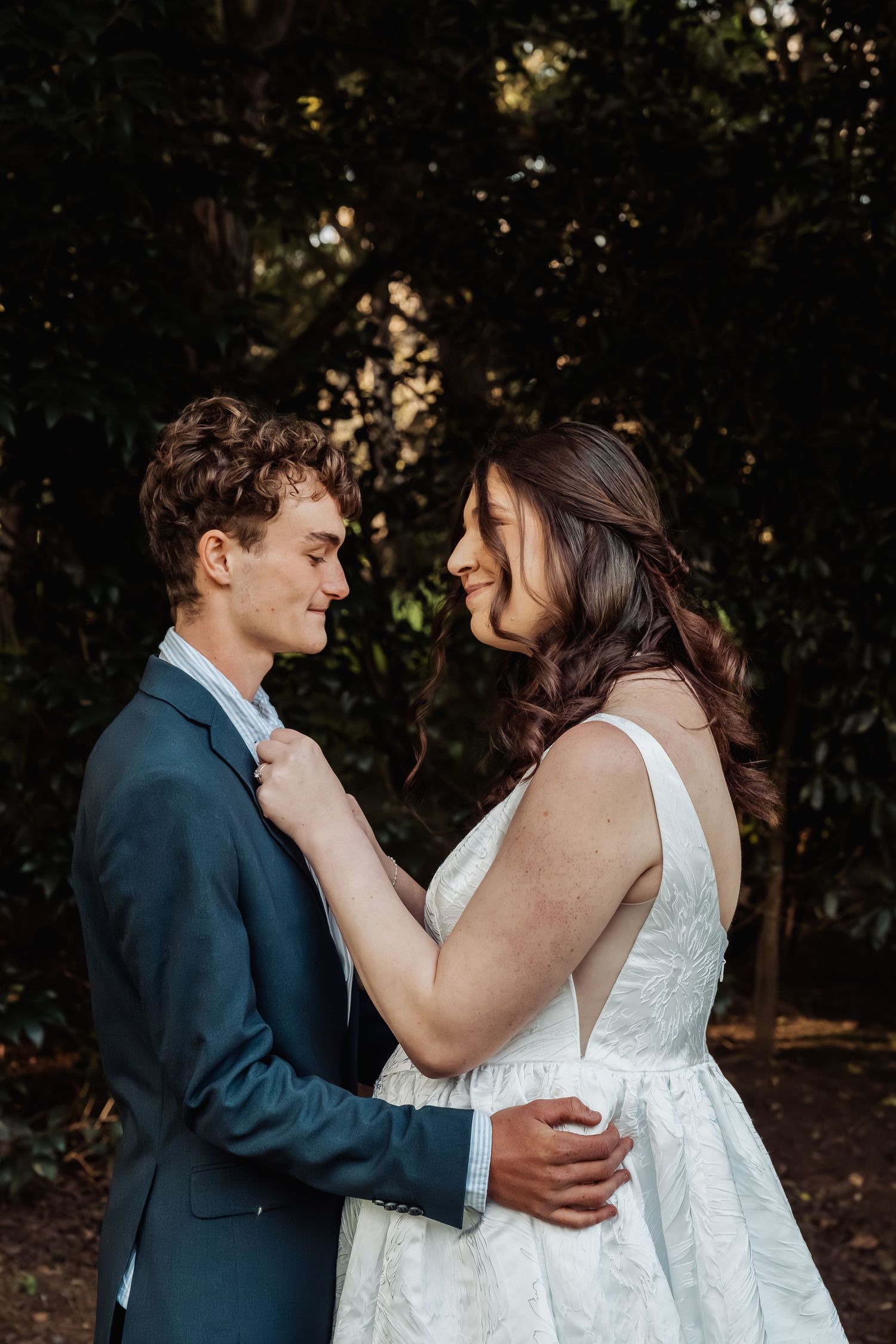 Bride and groom share an intimate moment surrounded by lush dark green foliage.