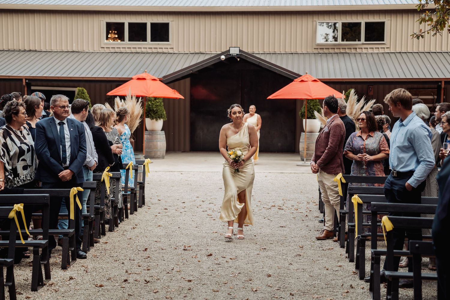 Bridesmaid walks down outdoor wedding aisle between seated guests at rustic barn venue with orange umbrellas.