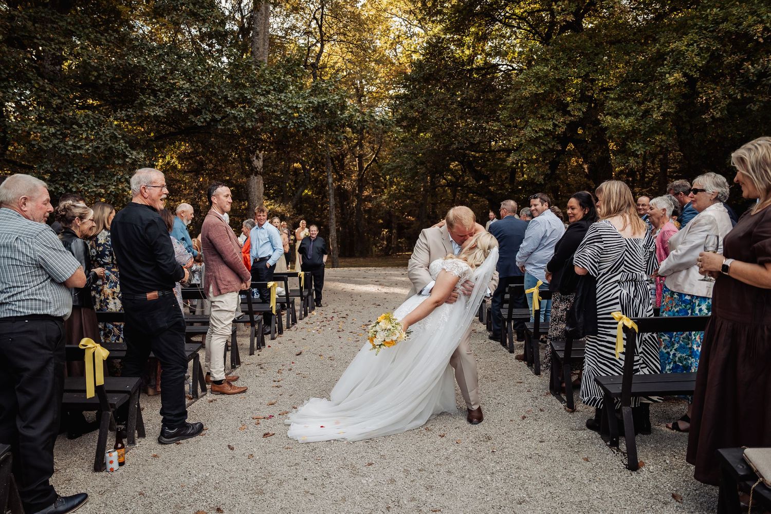 Groom dips bride in romantic kiss during outdoor wedding ceremony among guests.