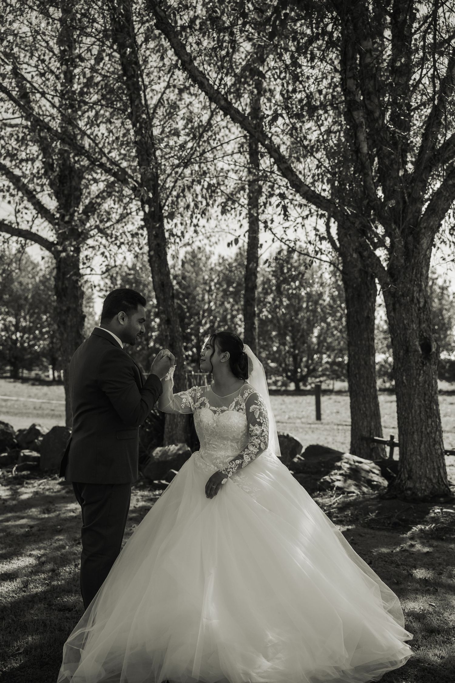 Black and white photo of bride in lace ball gown and groom holding hands among bare trees.