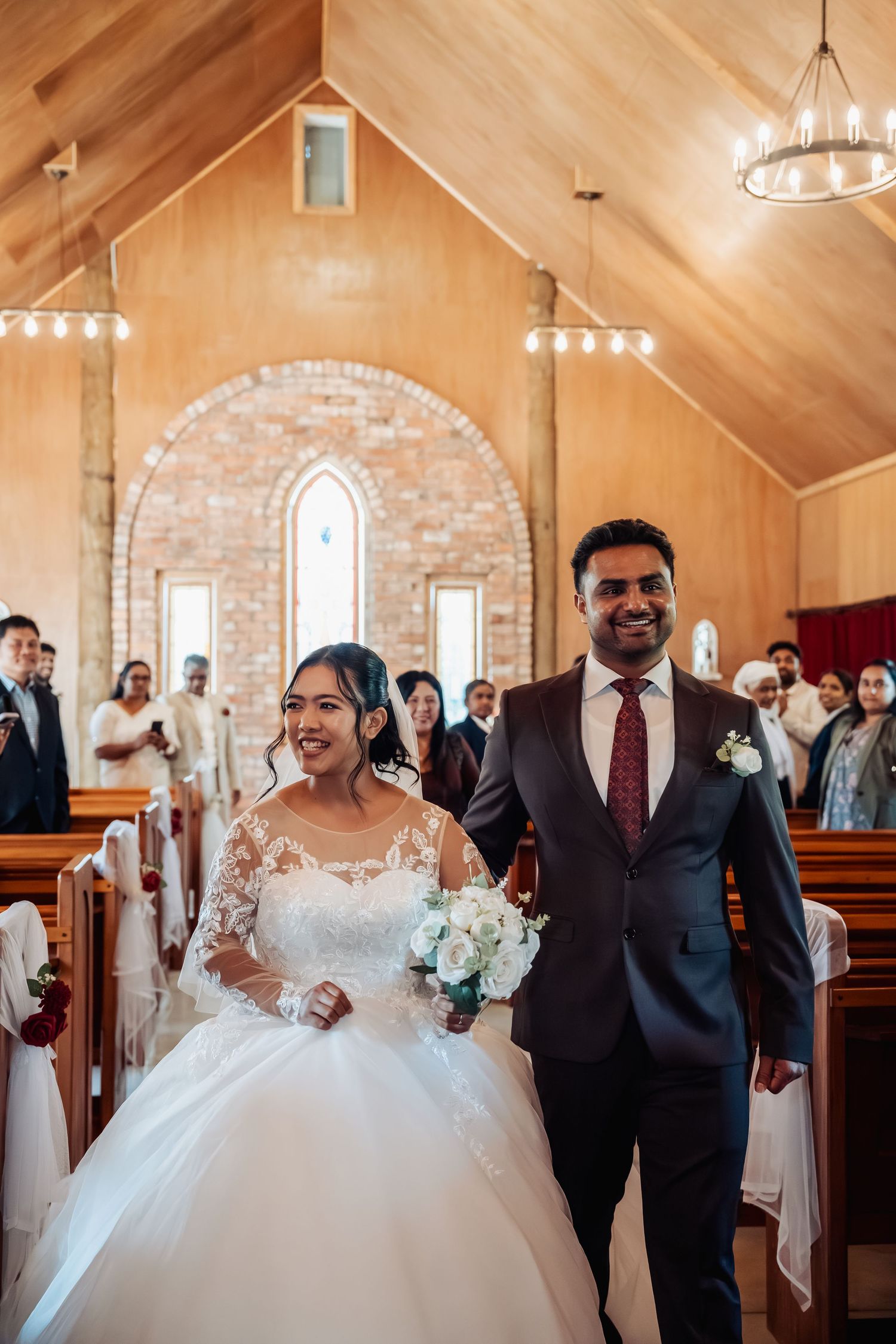 Smiling bride and groom walking down church aisle after wedding ceremony in wooden chapel.