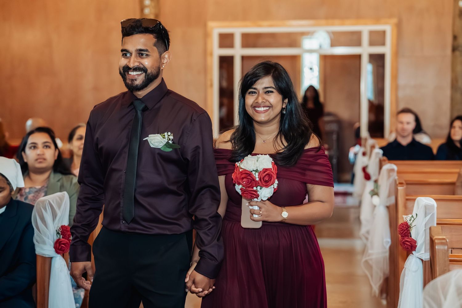 Couple in burgundy wedding attire walking down church aisle, man in dark shirt with boutonniere, woman holding red rose bouquet.