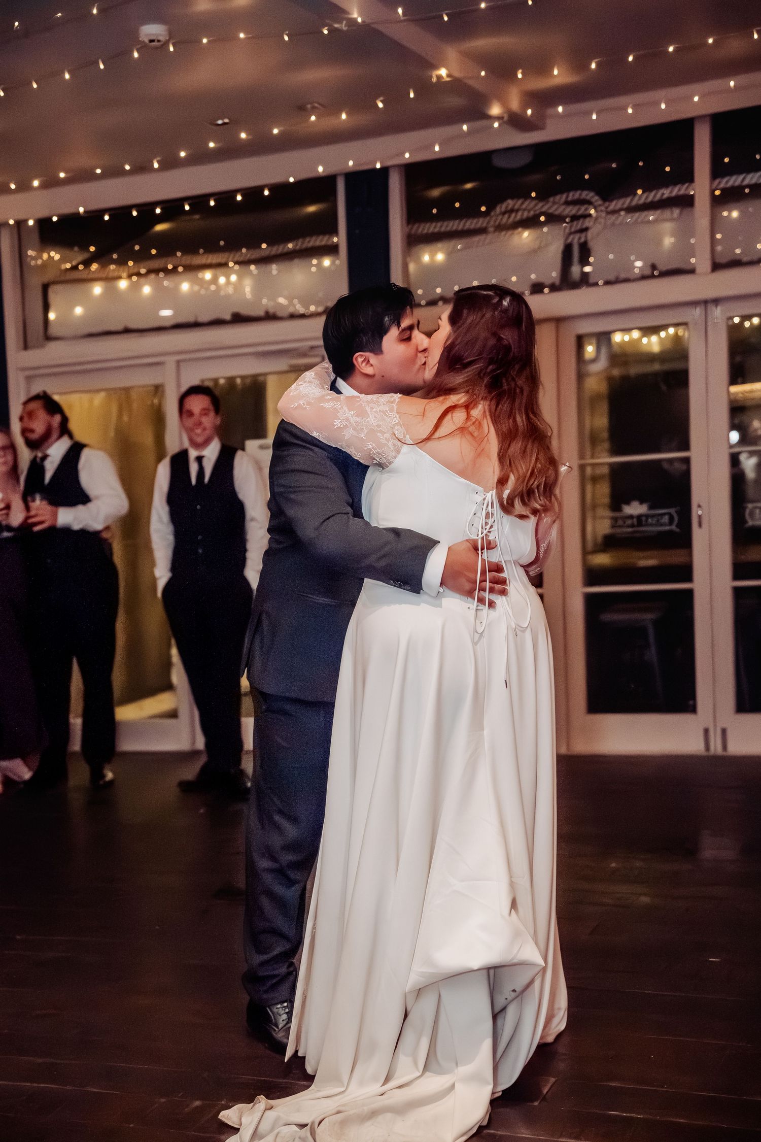 Couple shares romantic first dance as newlyweds under twinkling string lights at indoor wedding reception venue.