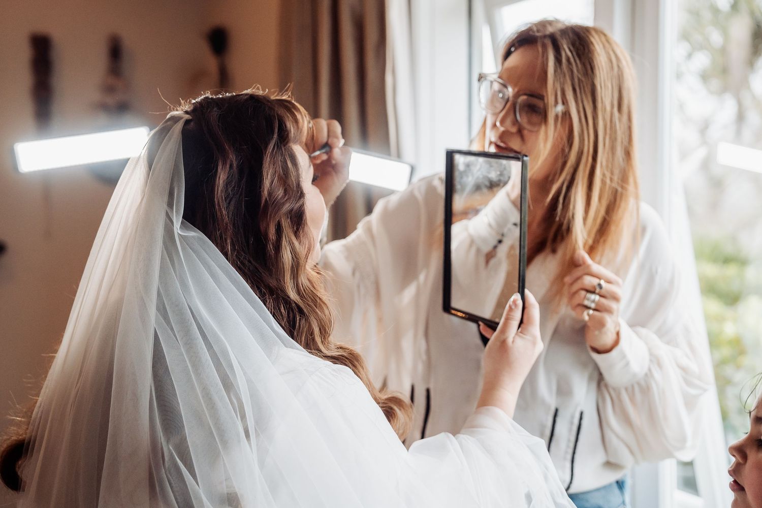 A bride getting her makeup done while holding up a mirror during wedding preparations.