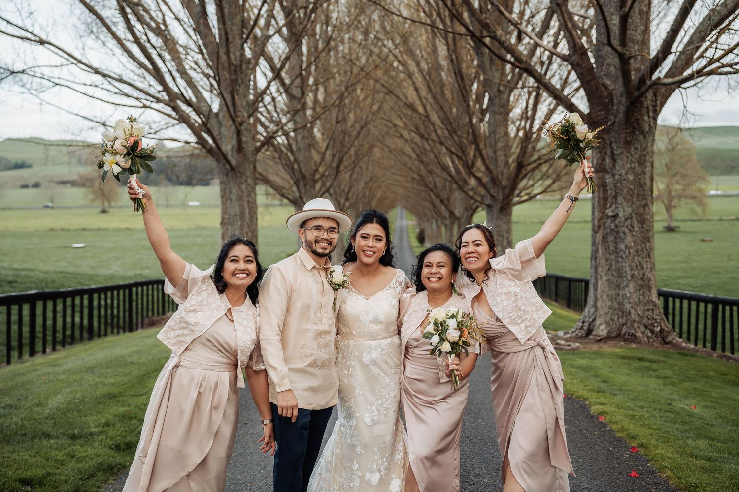 Wedding party in neutral tones poses beneath large trees on a grassy hillside.