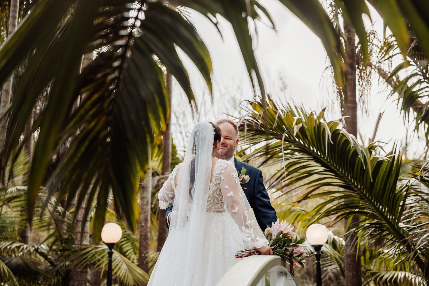 Wedding couple poses beneath tropical palm trees in an elegant garden setting.