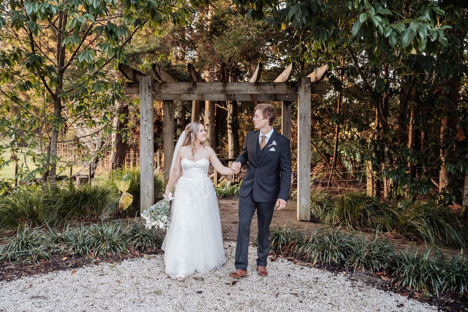A couple stands under a wooden garden arbor surrounded by lush greenery and trees on a gravel path.