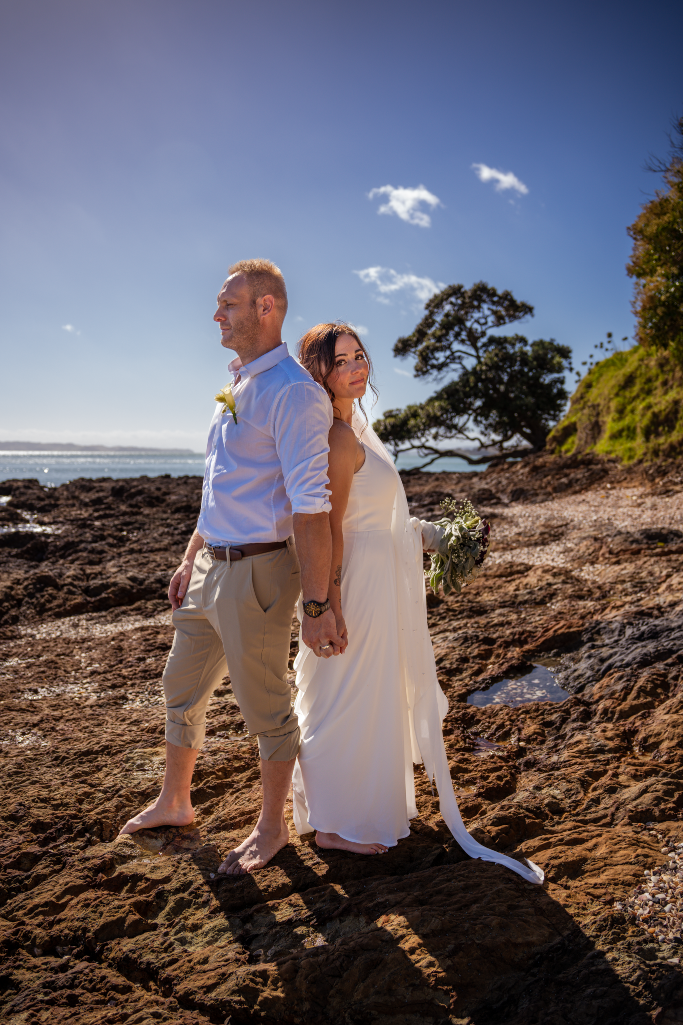 A couple in wedding attire stands together on coastal rocks with ocean views.