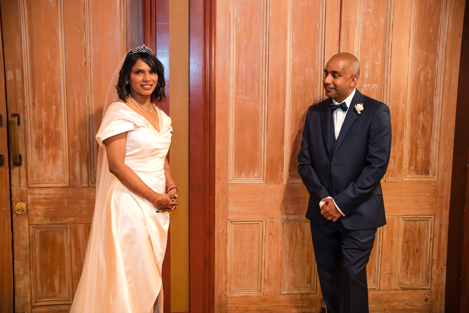A couple in wedding attire pose formally against wooden paneled walls.