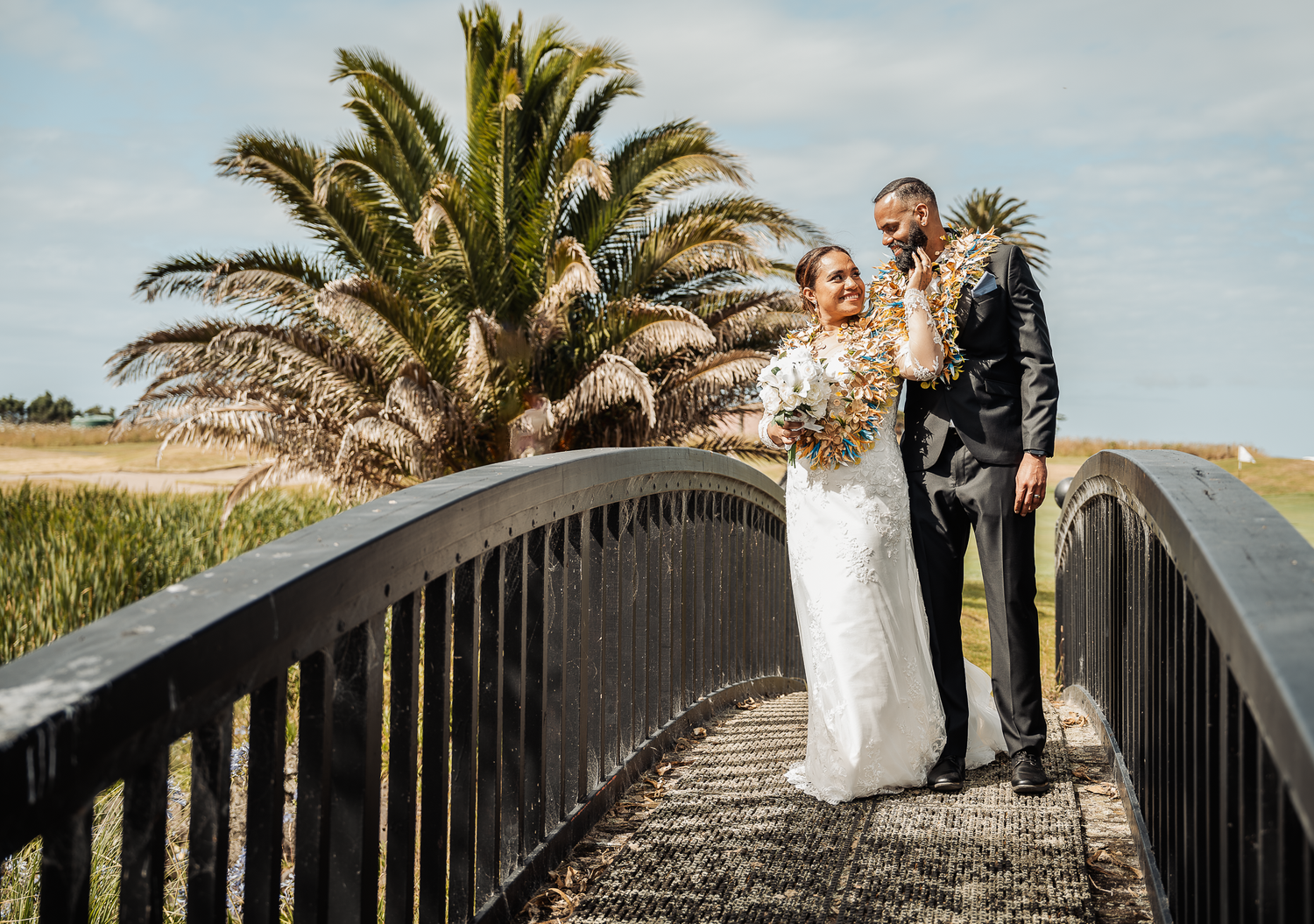 Wedding couple poses on wooden walkway with palm tree framed against cloudy sky.