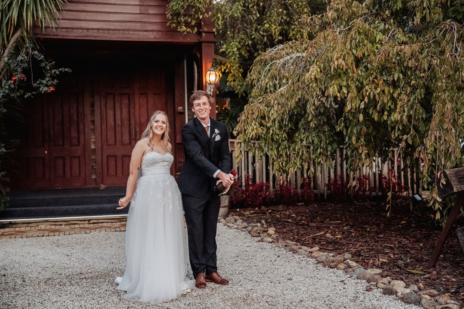 Two people pose together in formal wedding attire in front of a red wooden barn and lush greenery.