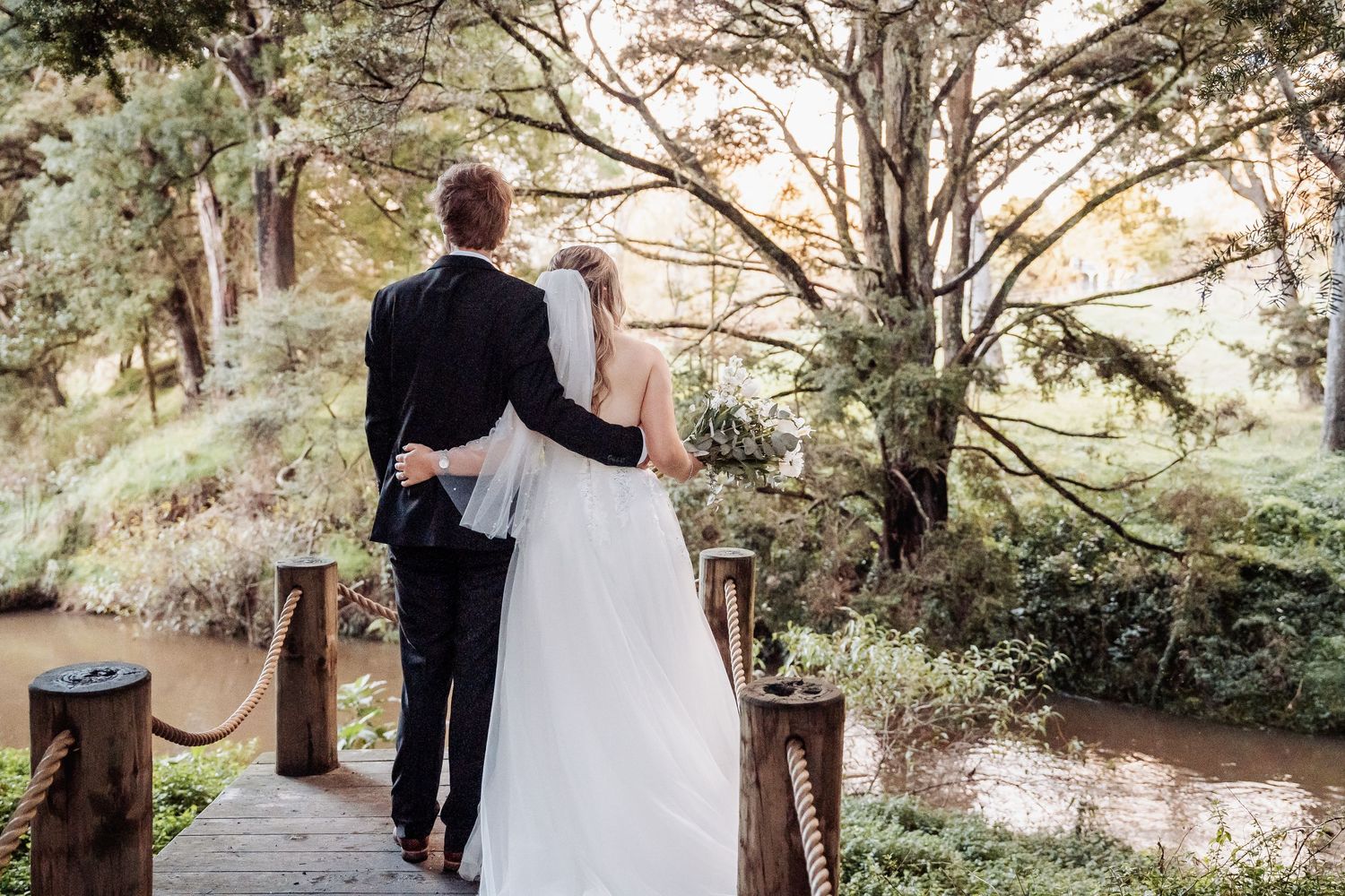 A romantic couple stands together on a wooden bridge during sunset in a lush garden setting.