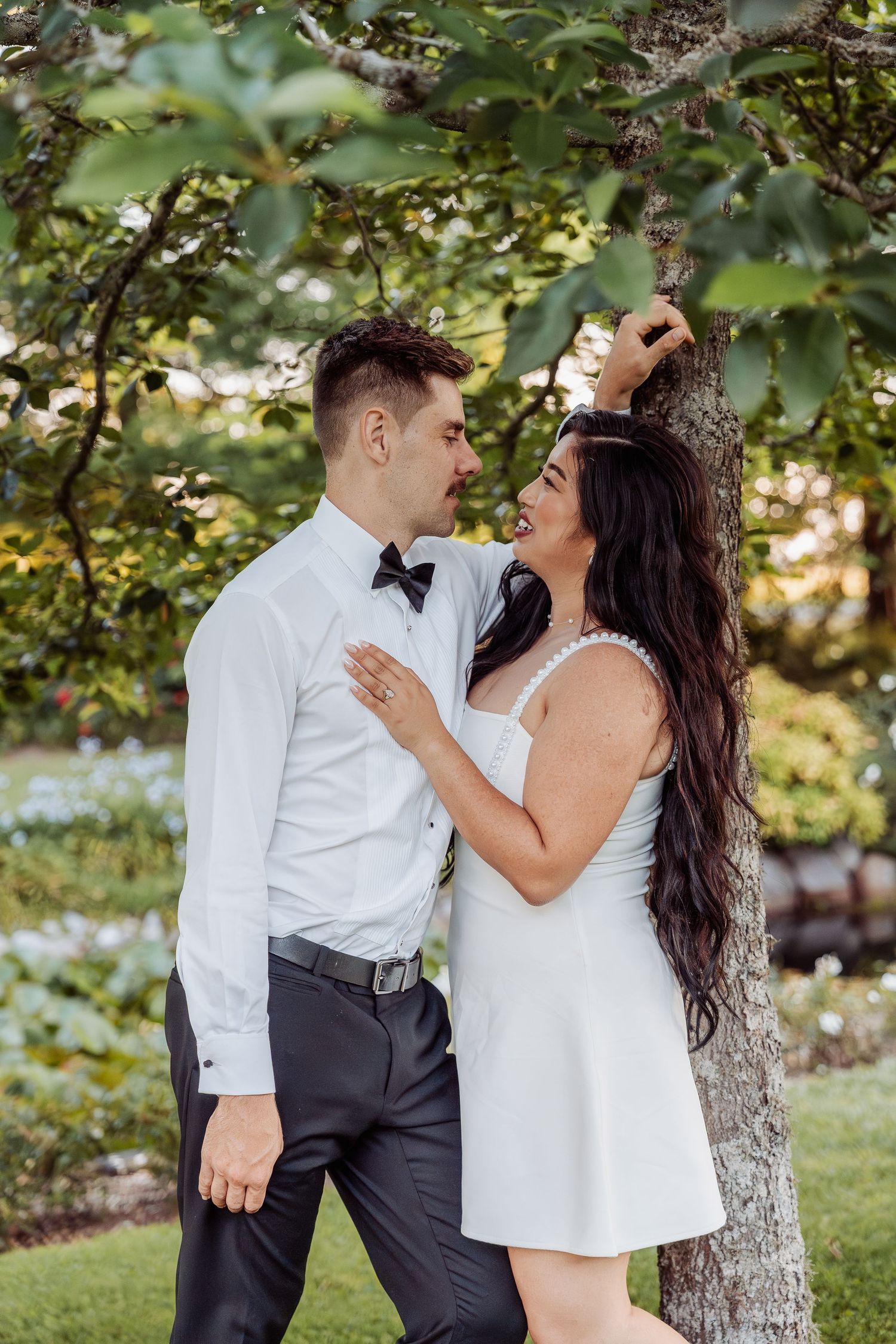 Laughing bride in white mini dress leaning against tree with groom in garden.