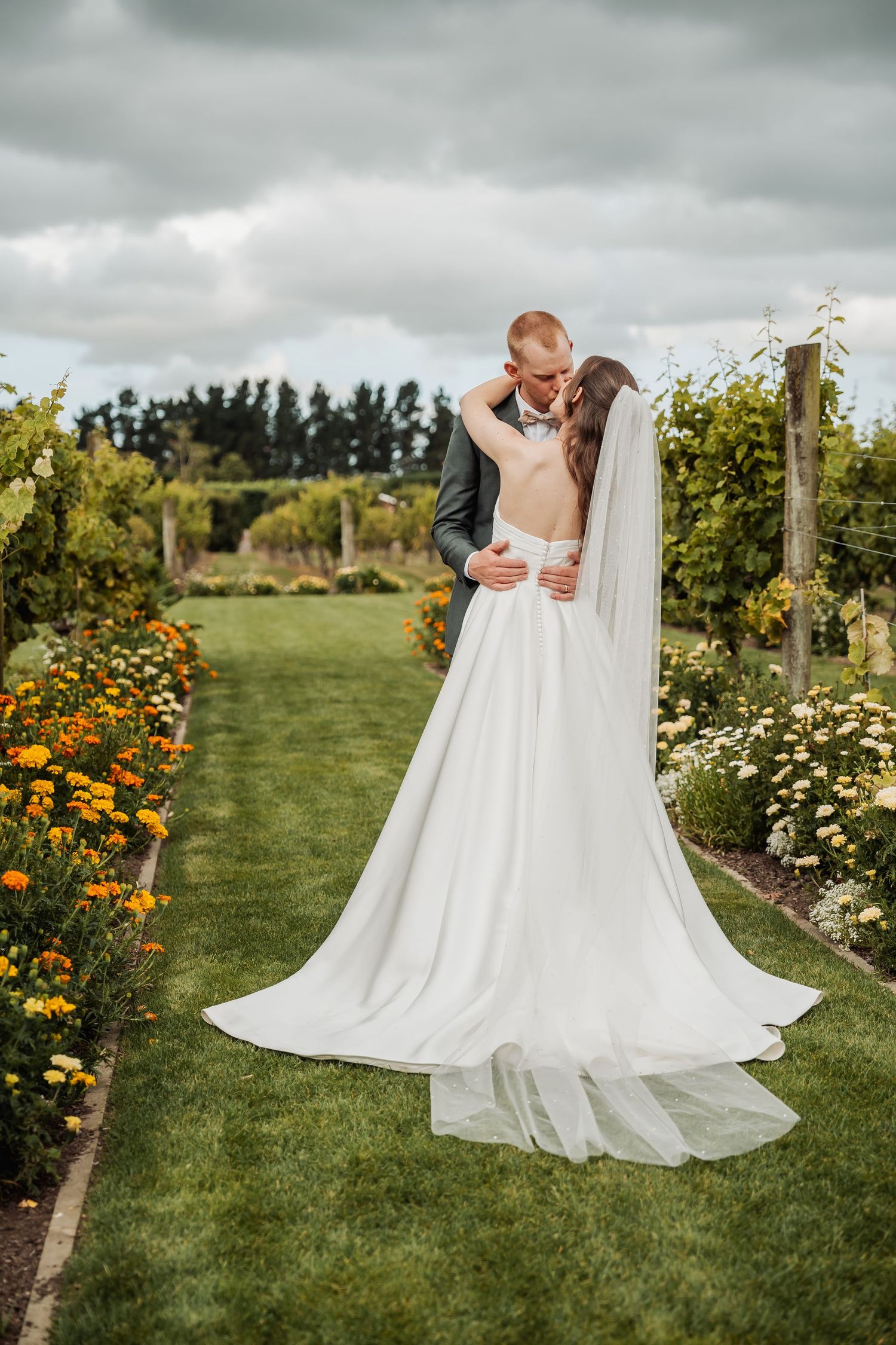 Bride and groom share a passionate kiss surrounded by colorful flower gardens in a scenic vineyard.