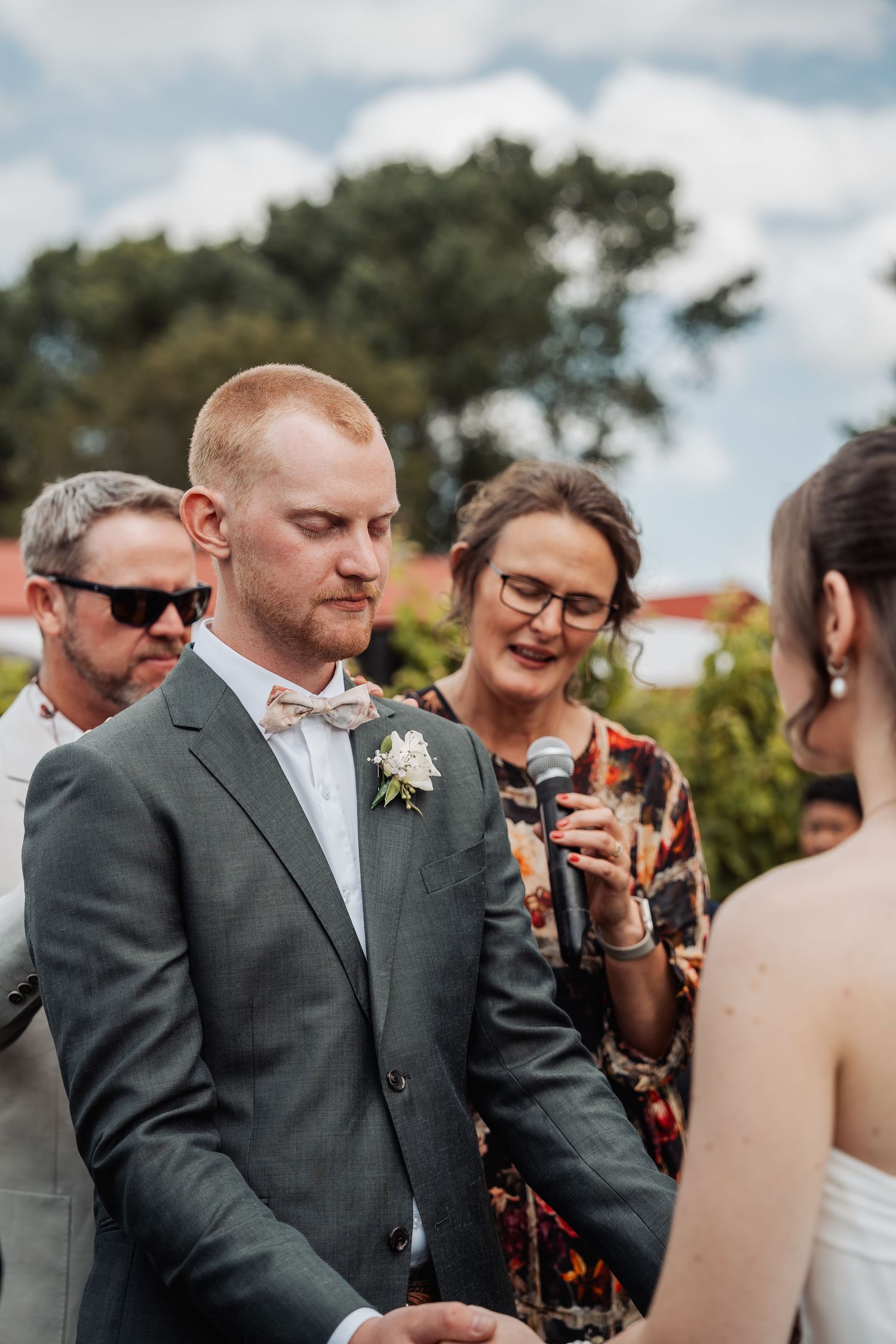Groom in grey suit with bow tie listening during outdoor wedding vows while officiant holds a microphone.