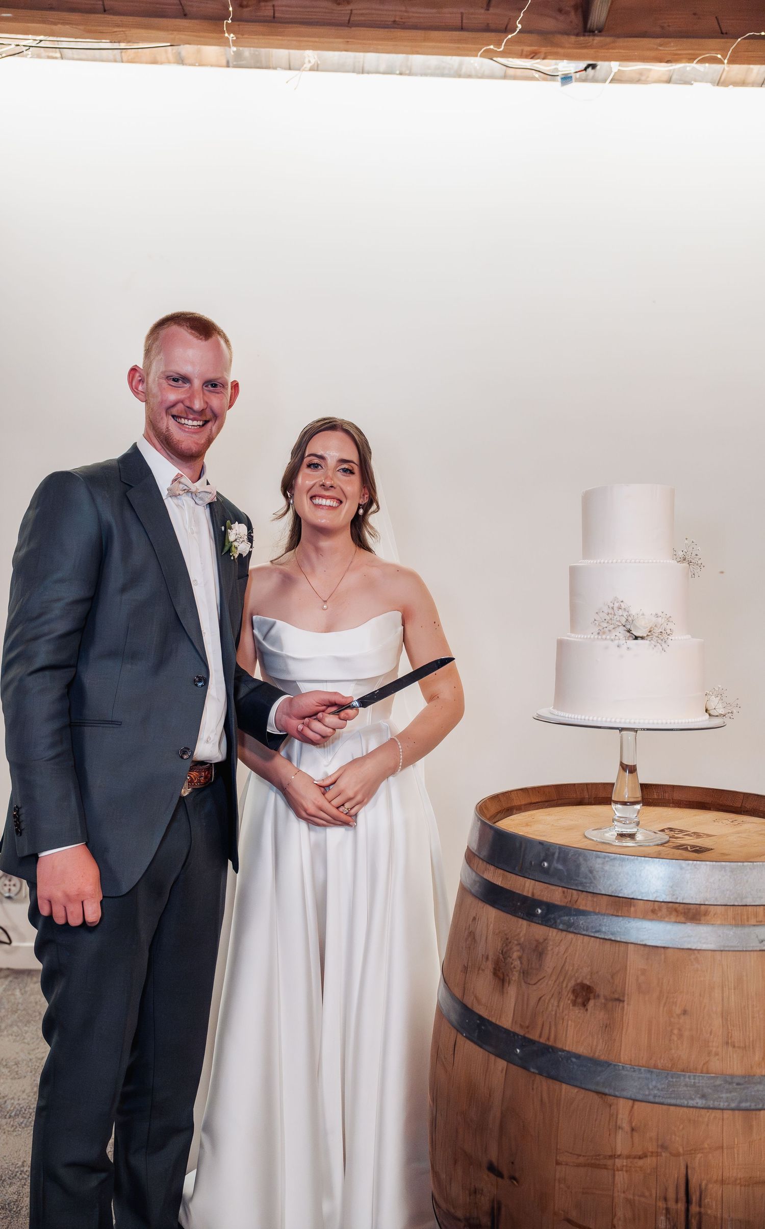 Smiling newlyweds in wedding attire pose together beside their three-tier white cake displayed on a wooden barrel.