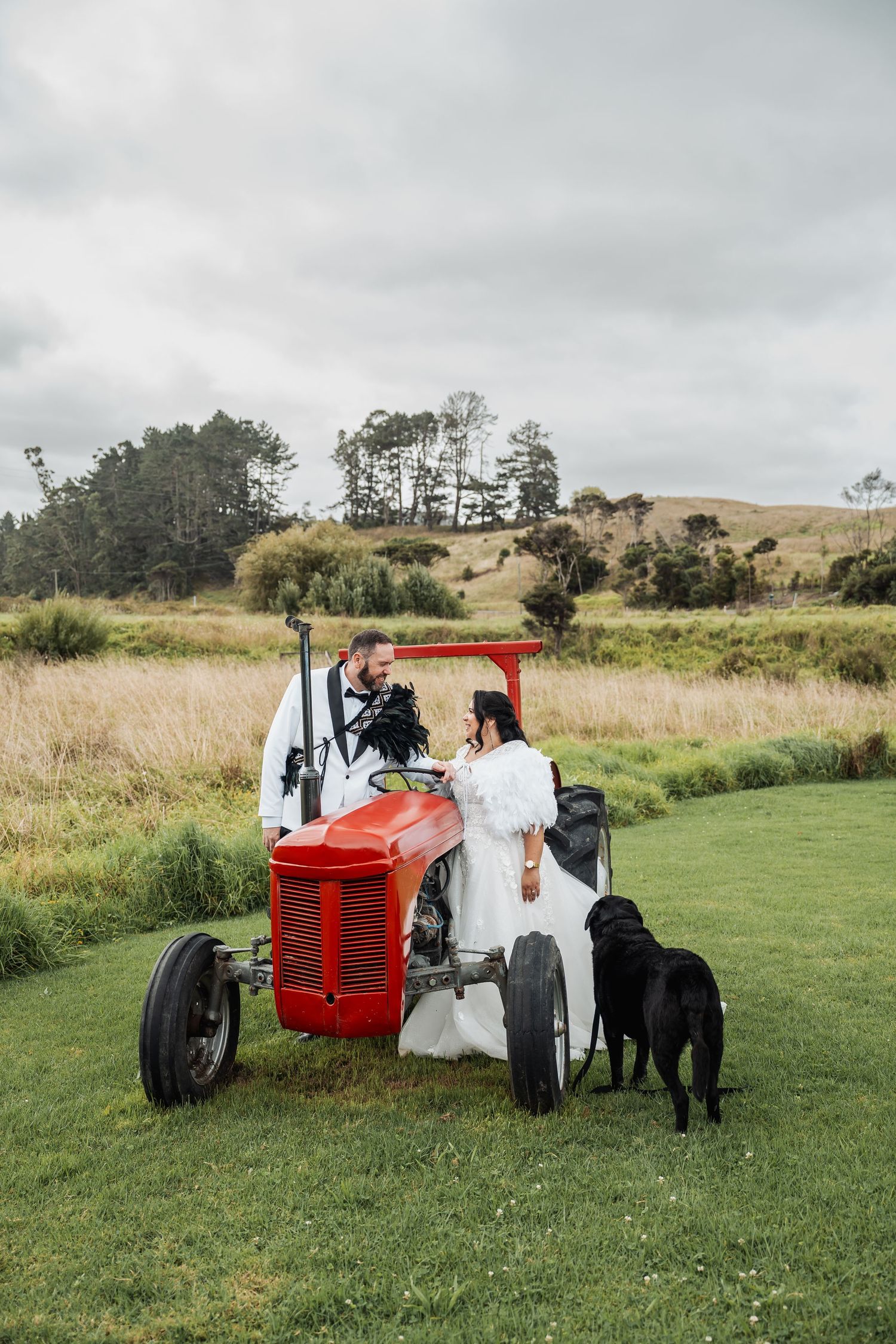 Bride and groom share a romantic moment by a red tractor with a black dog nearby.