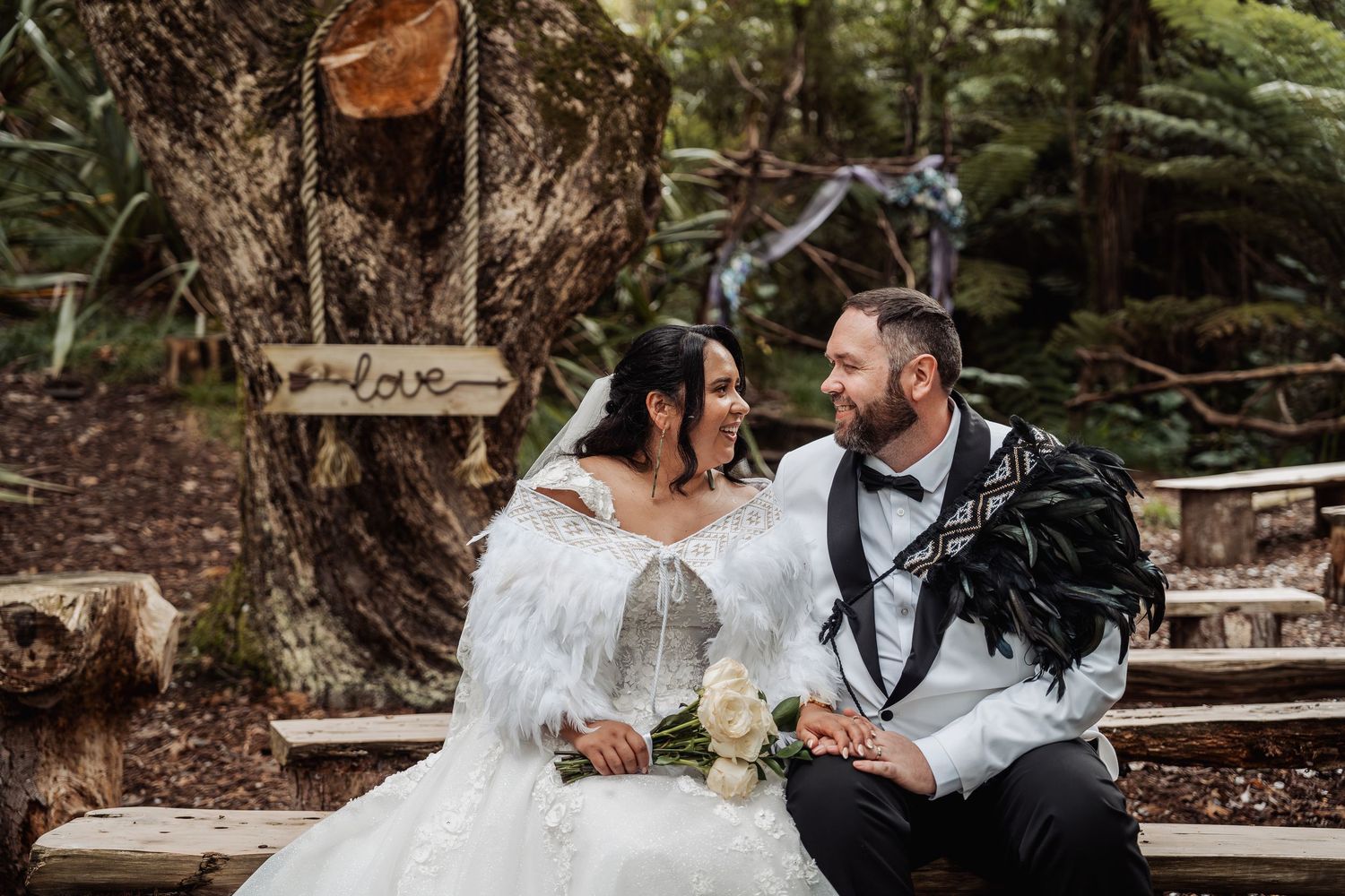 Bride and groom sitting together on a wooden bench in a forest wedding setting, smiling at each other.
