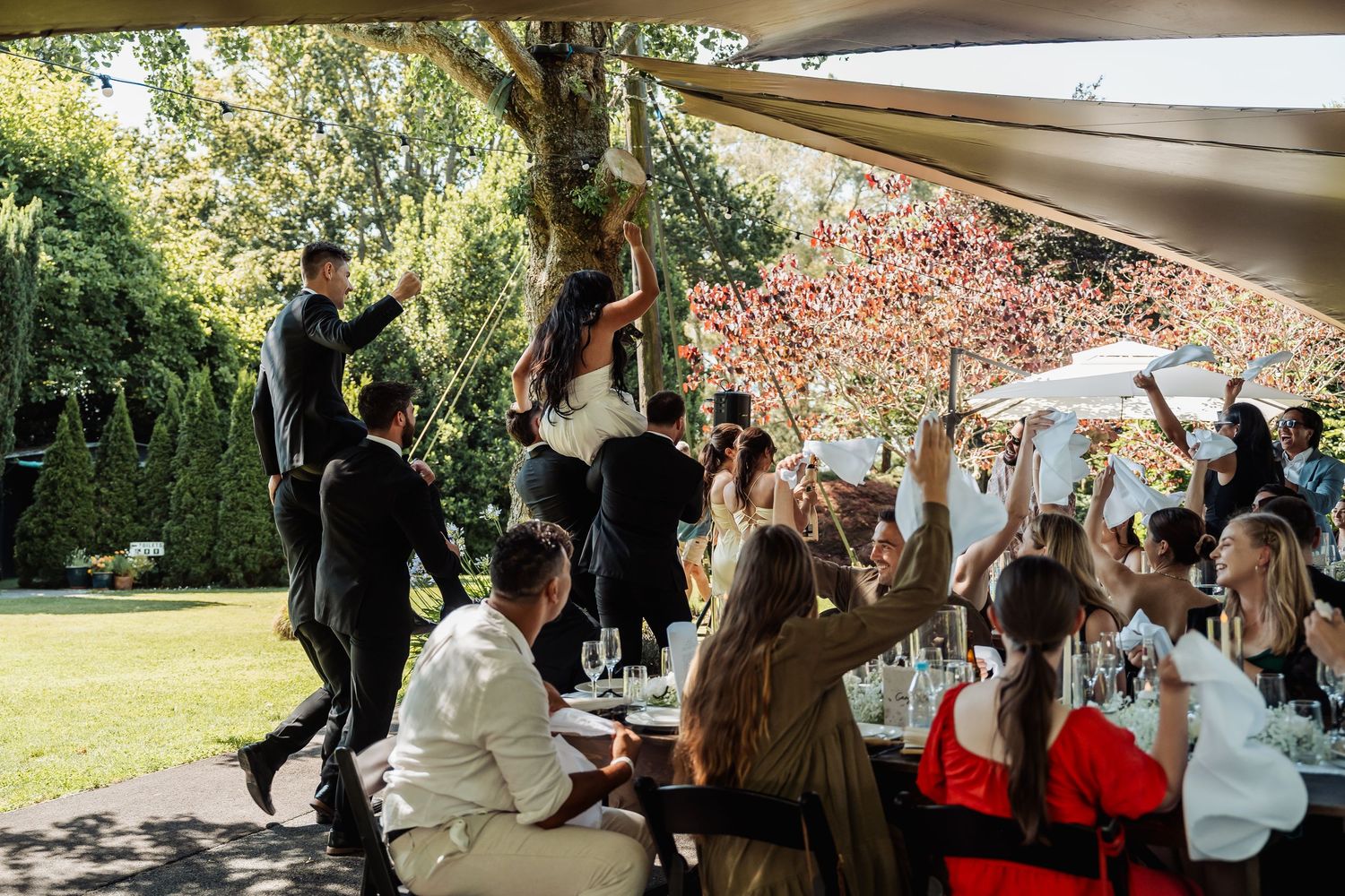 Outdoor wedding reception under a canopy with guests celebrating, raising glasses, and dancing in a lush garden setting.