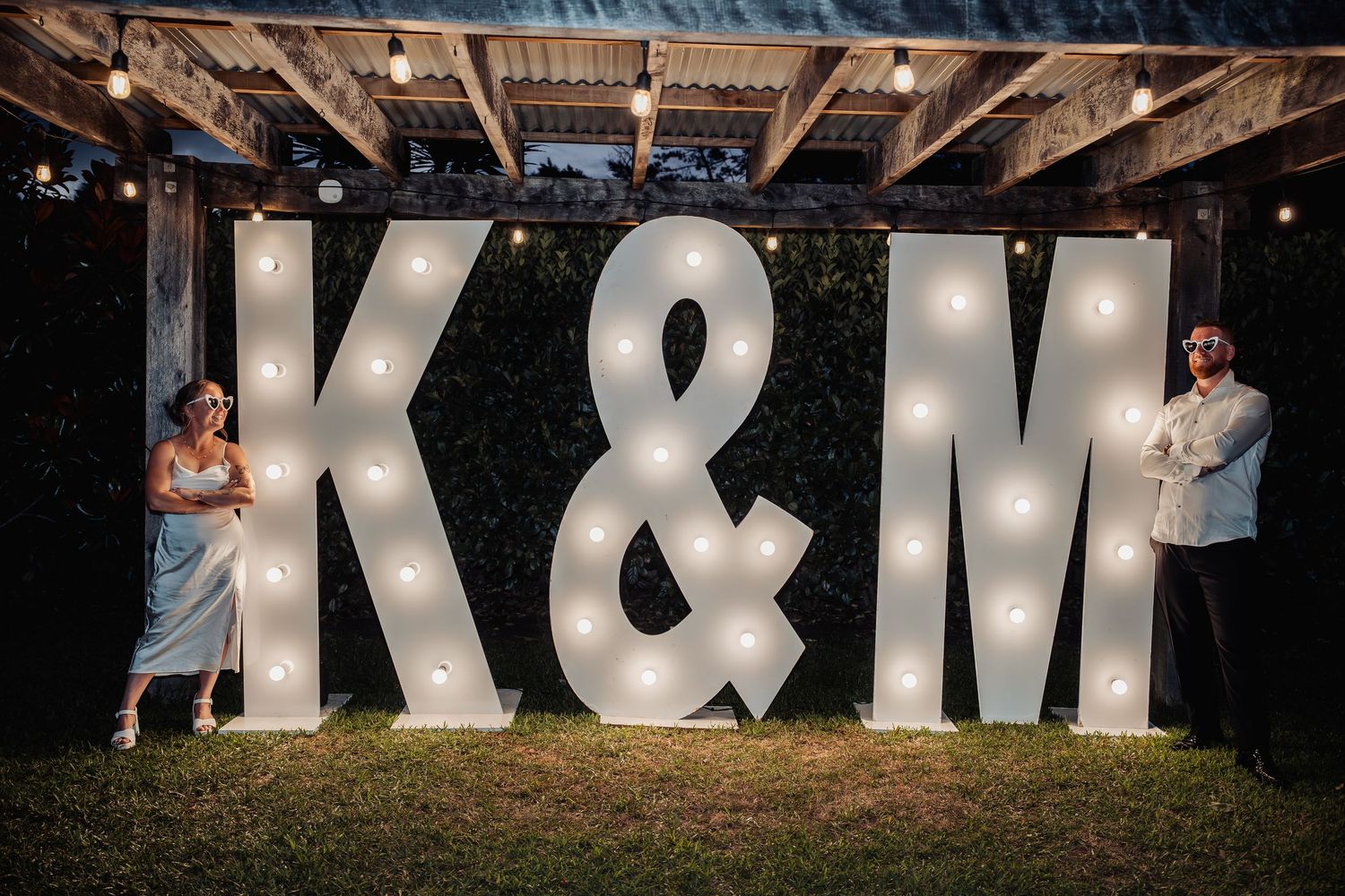 Couple posing beside illuminated marquee letters K&M at outdoor evening wedding reception.