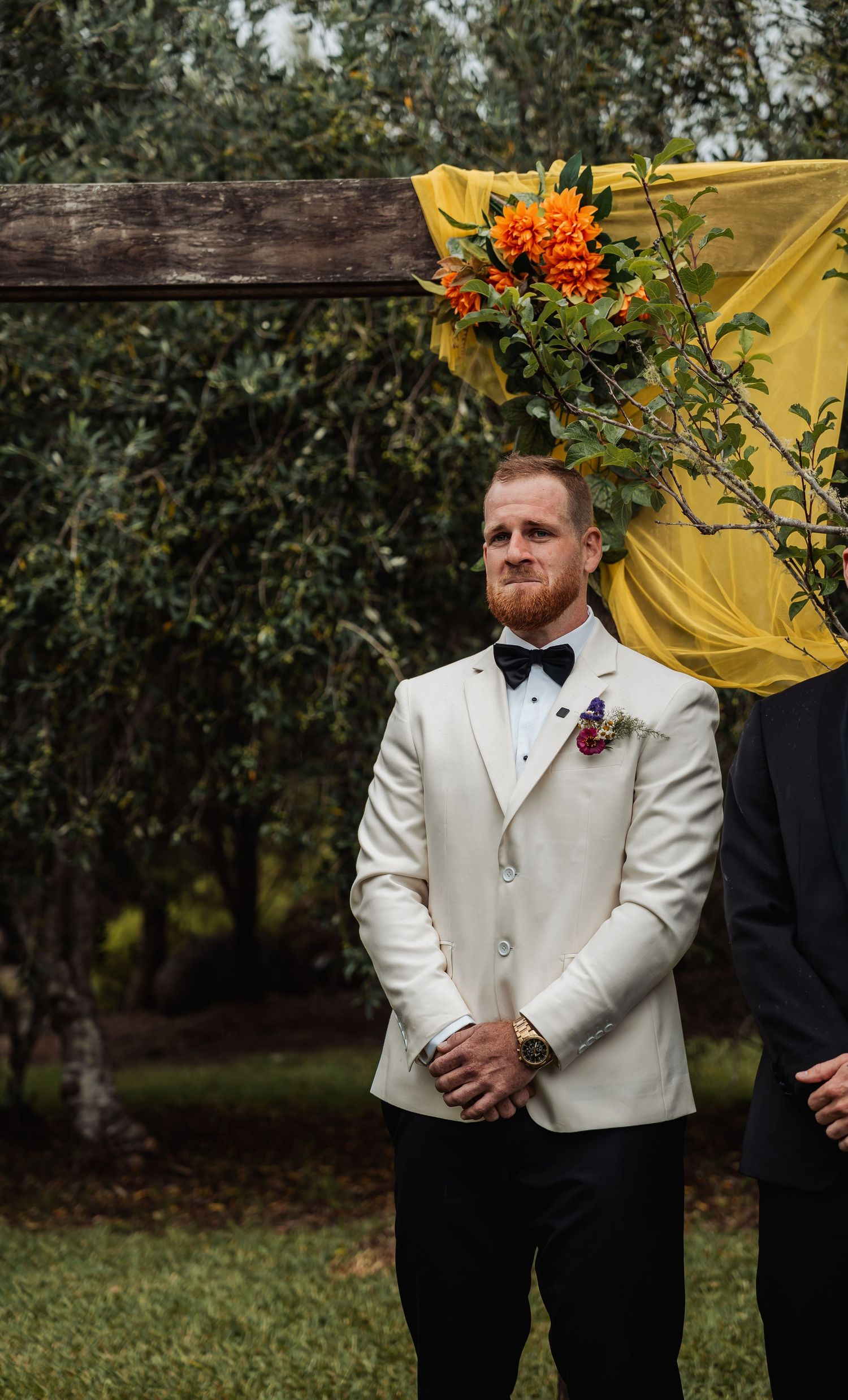 Groom in white tuxedo jacket with bow tie standing at outdoor wedding ceremony altar.