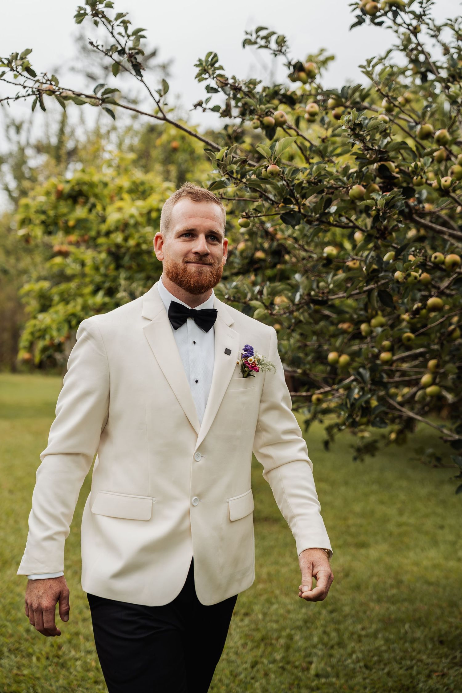 Groom in cream tuxedo jacket and black bow tie standing near an apple tree outdoors.