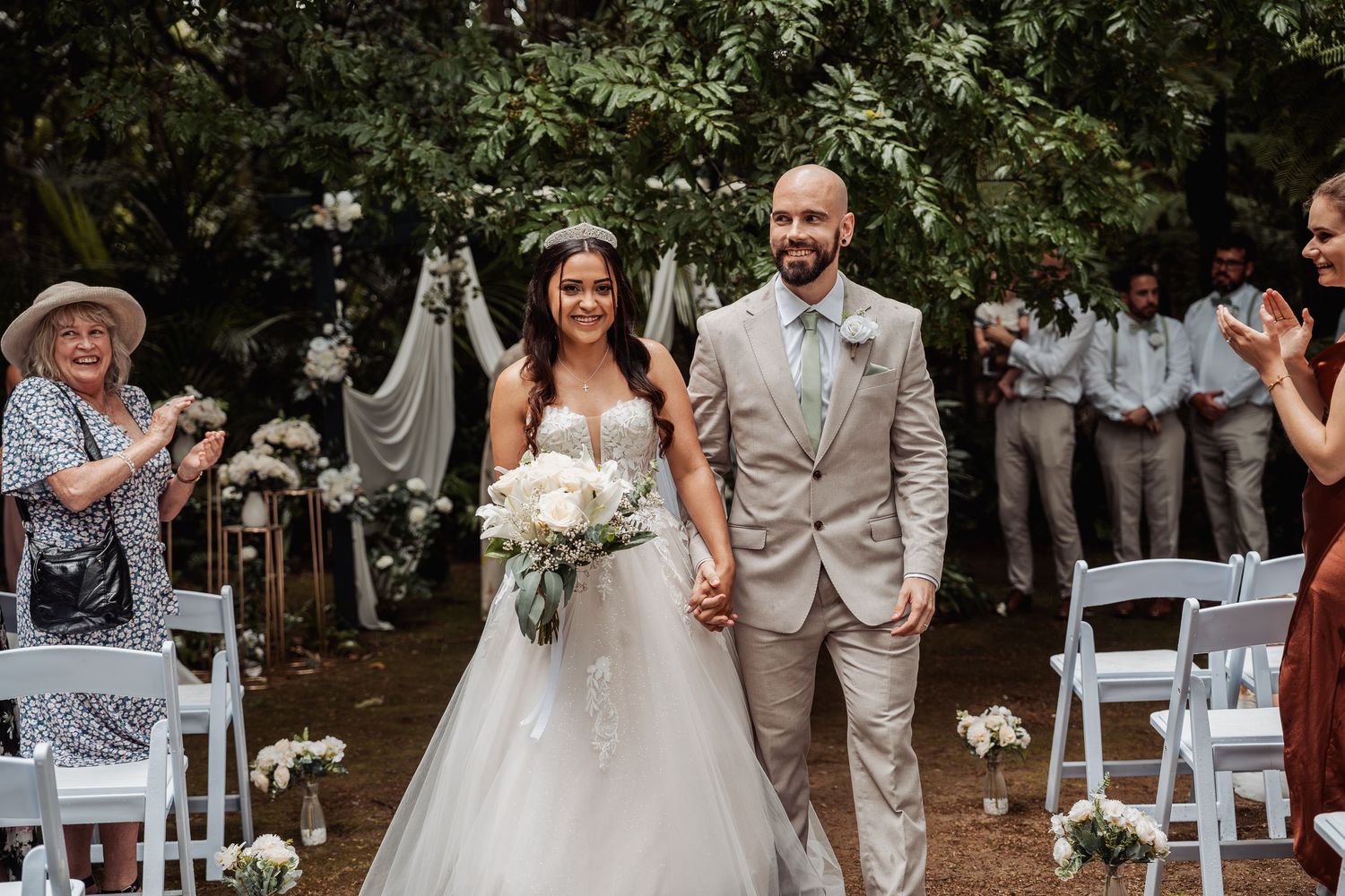 Newlywed couple walks down the aisle hand-in-hand at an outdoor garden wedding ceremony with guests applauding.