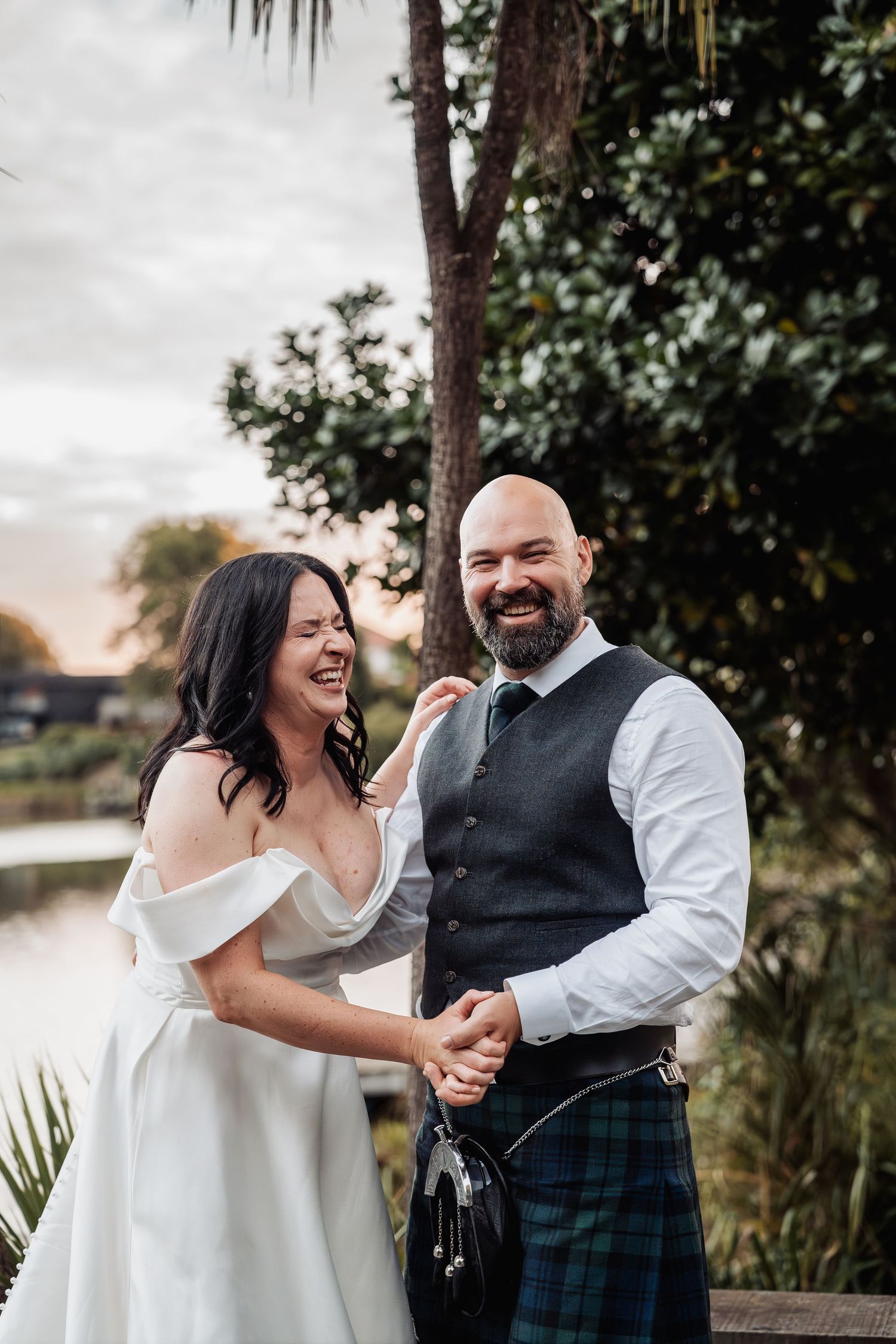 Laughing bride and groom in Scottish kilt share a joyful moment by a lake.