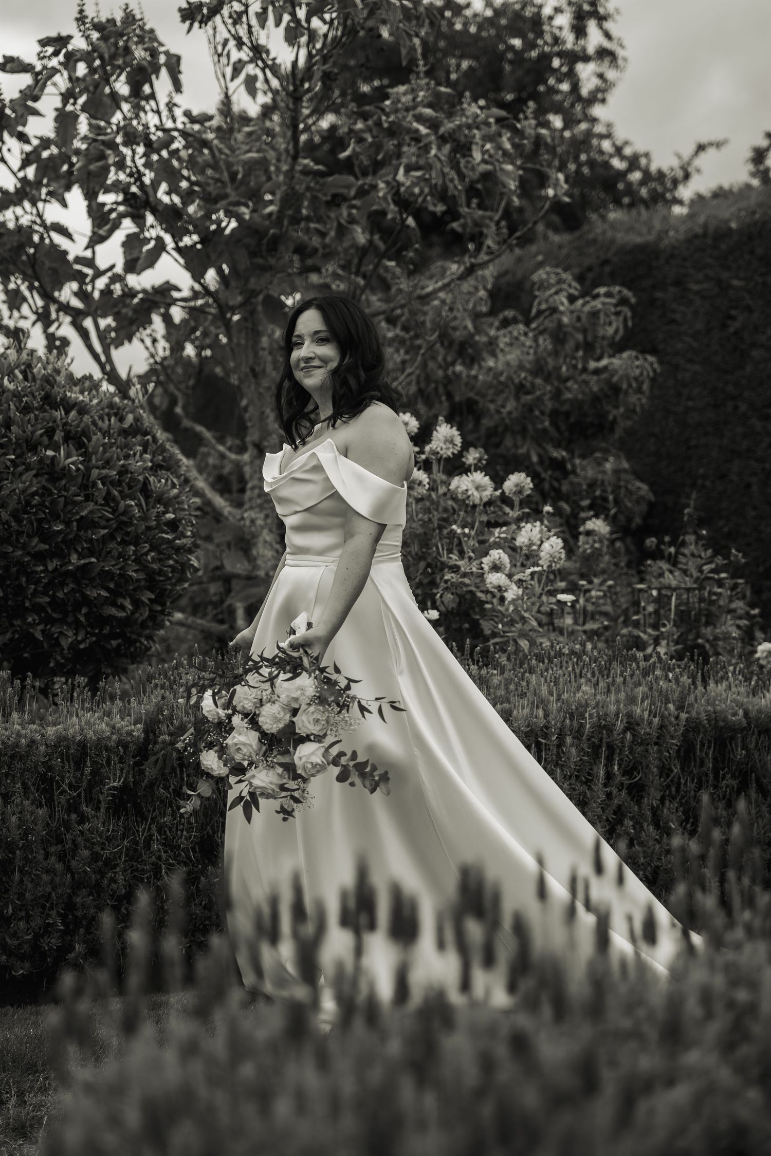 Black and white portrait of smiling bride in off-shoulder satin gown holding a bouquet in a lush garden.
