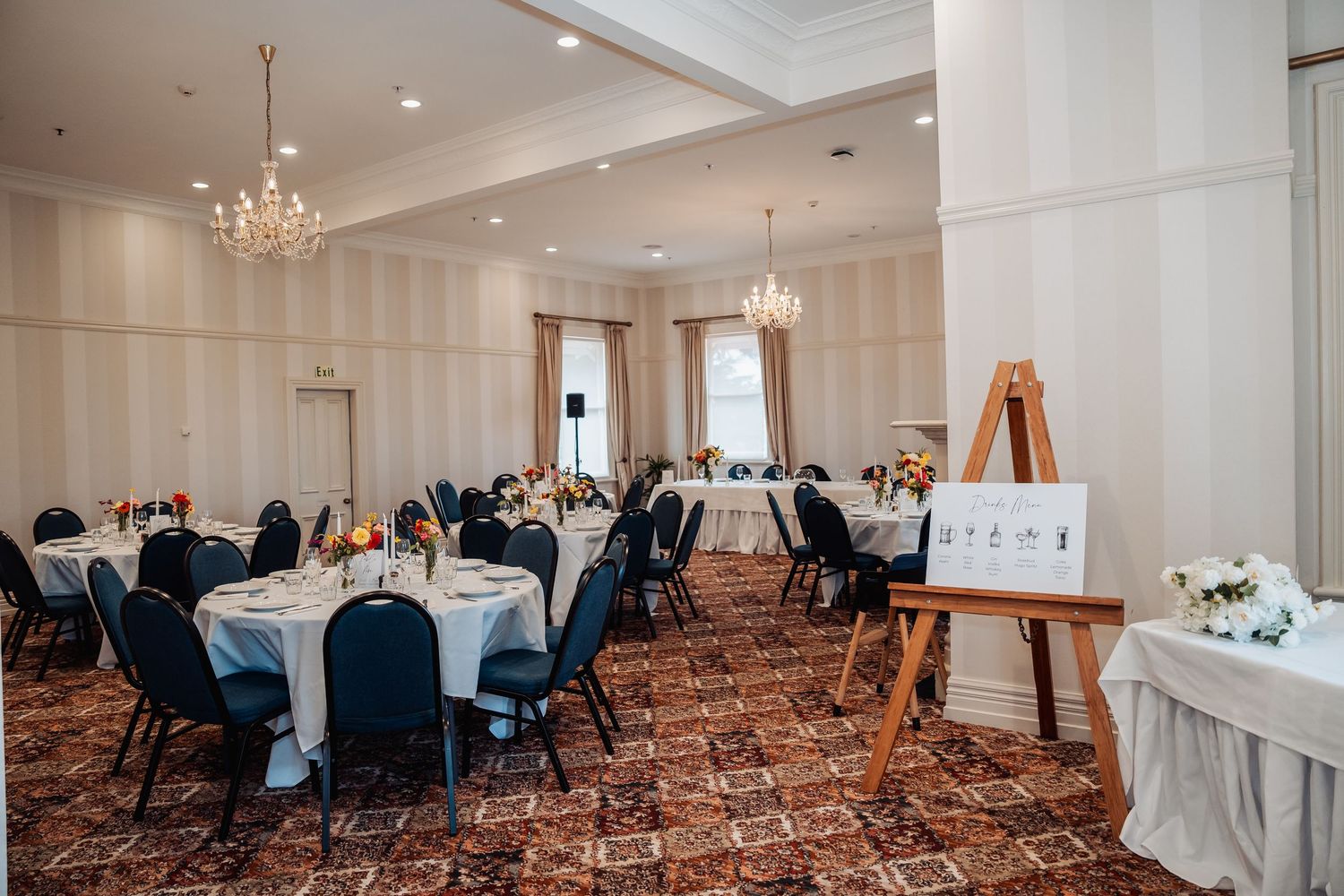 Elegant banquet hall with round tables, black chairs, chandelier lighting, and an easel display, set for a wedding reception.