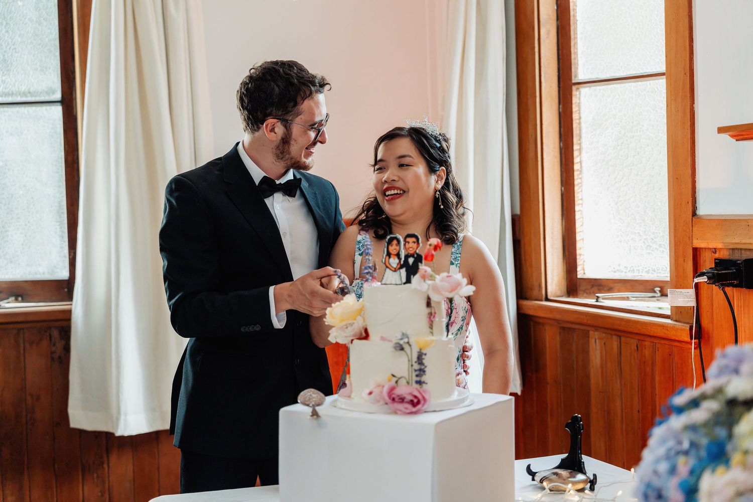 Smiling bride and groom cutting their decorated wedding cake together indoors at their wedding reception.