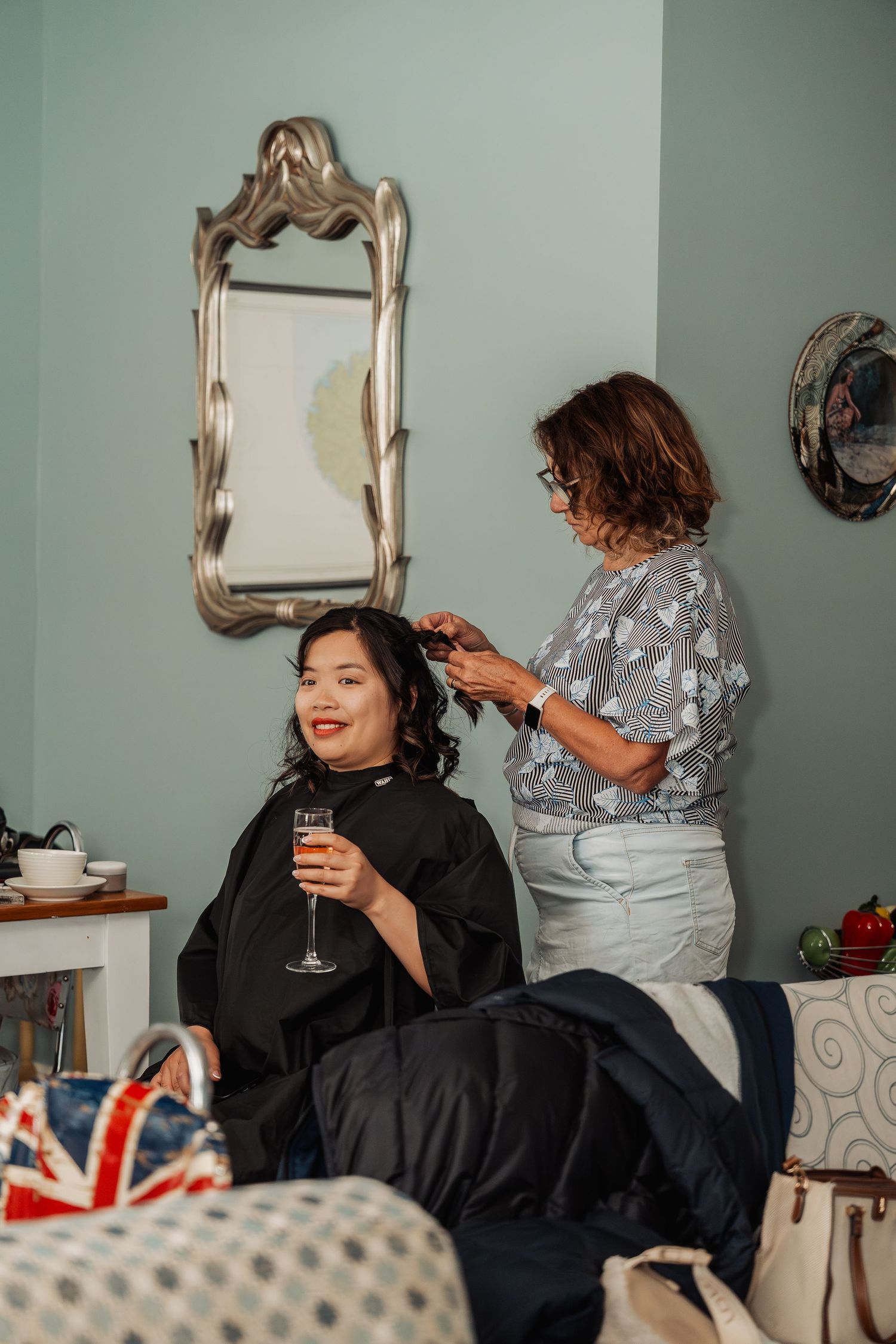 Bride smiling in salon chair holding champagne while hairstylist works on her curly hair before wedding.