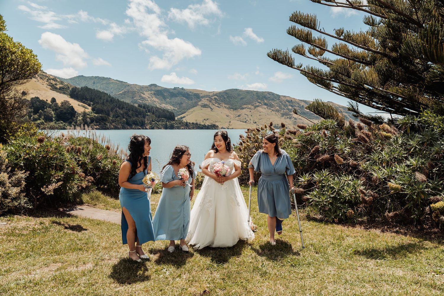 Bride in white gown walks with three bridesmaids in blue dresses by scenic lake and mountains.