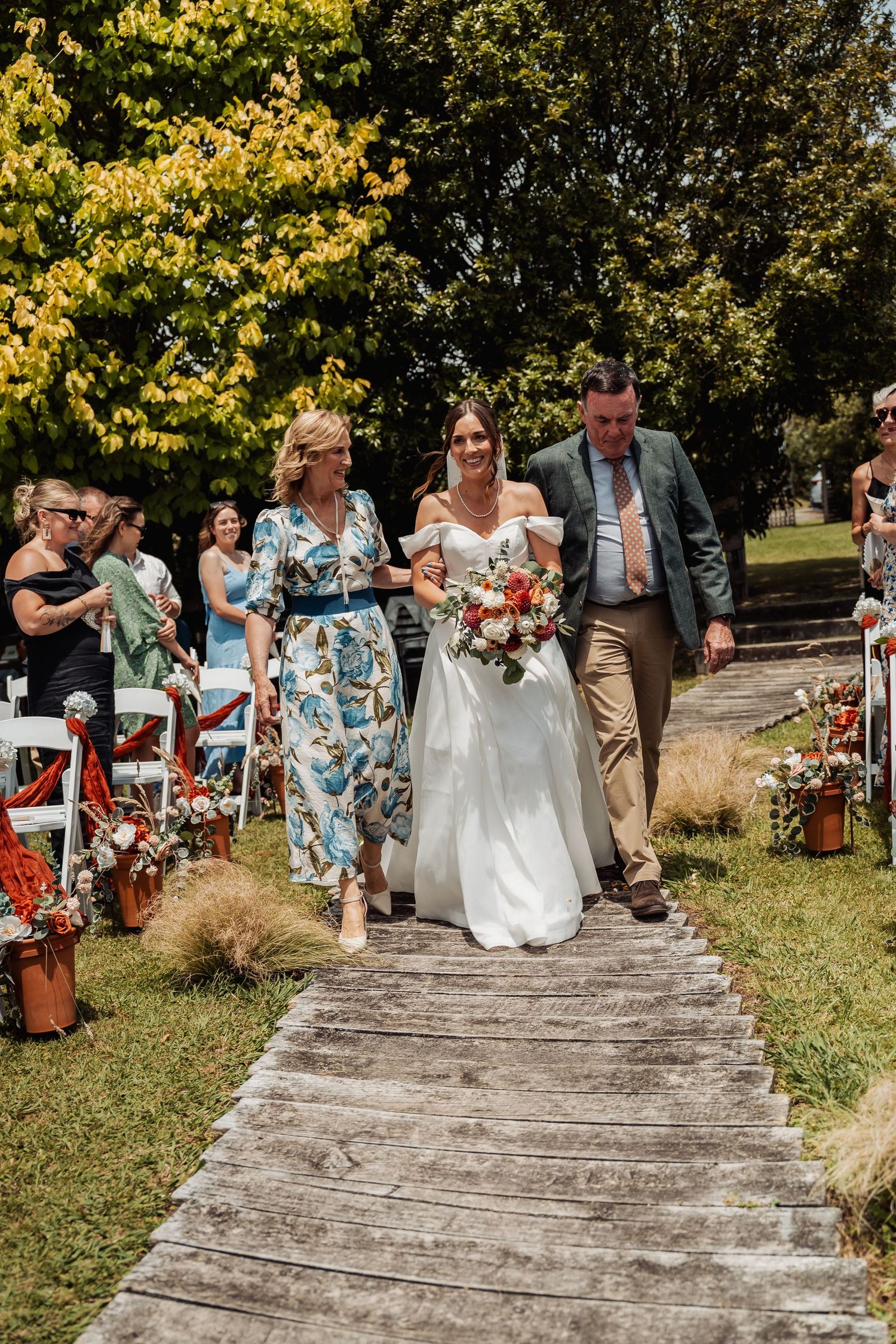 Bride walks down outdoor aisle escorted by parents at a sunny garden wedding ceremony.
