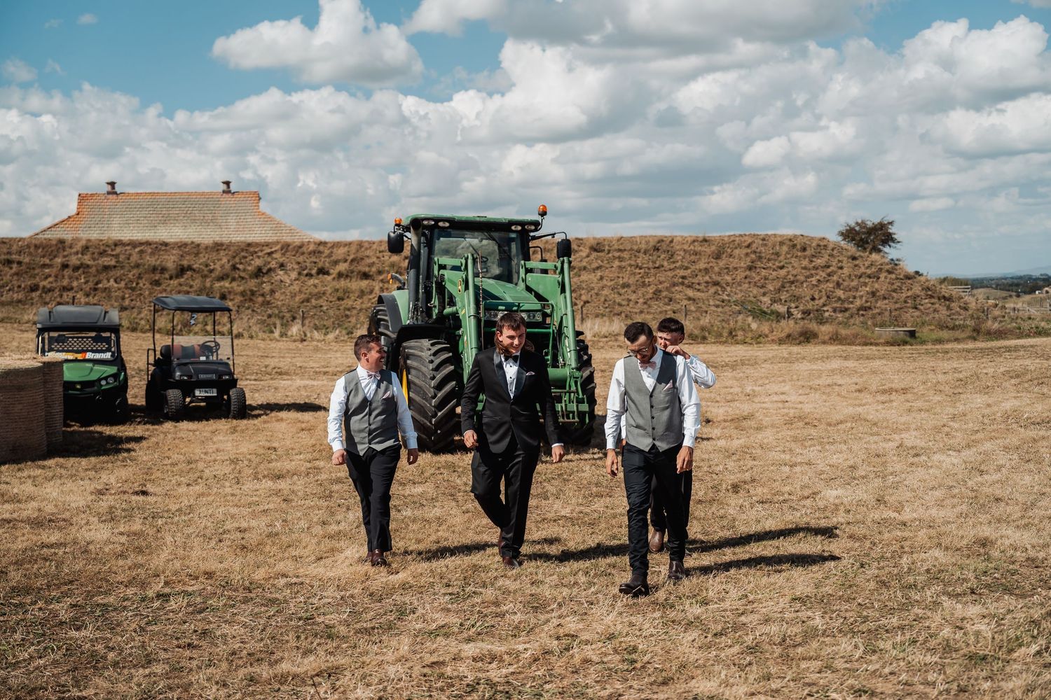 Three groomsmen walking confidently across a rustic farm field with a green tractor in the background.