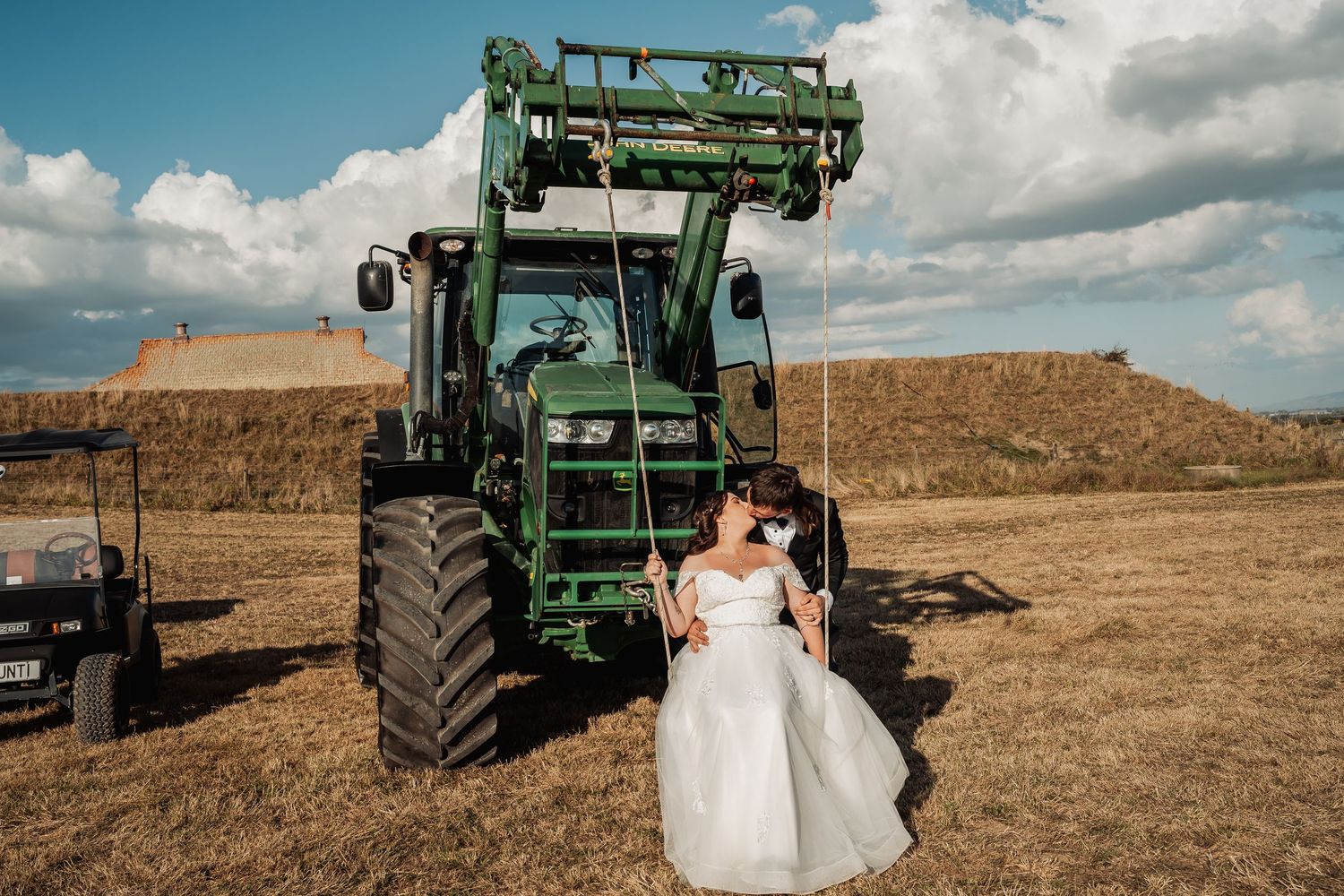 Bride in white wedding gown sitting on a swing attached to a large green tractor in a sunny field.