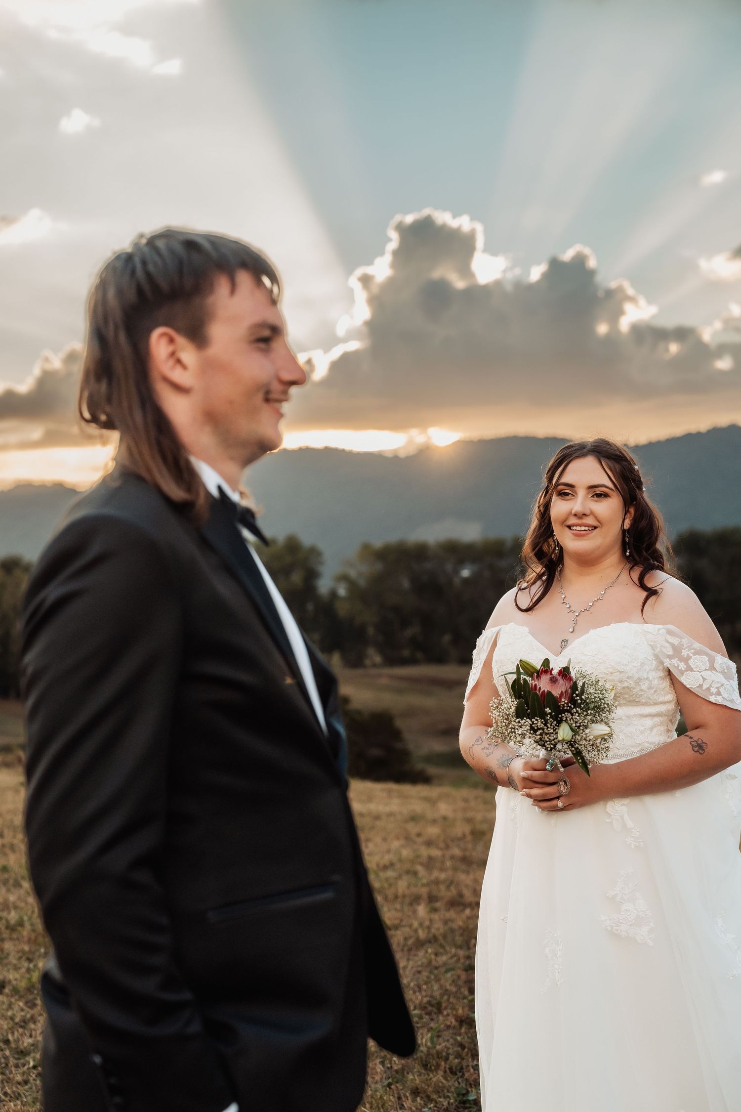 Bride in white gown holds bouquet as groom in tuxedo stands before stunning sunset backdrop.