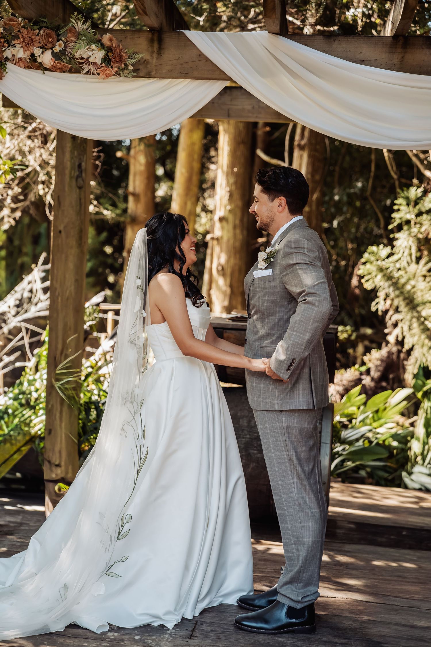 Smiling bride and groom hold hands at elegant outdoor woodland wedding ceremony under draped arbor.