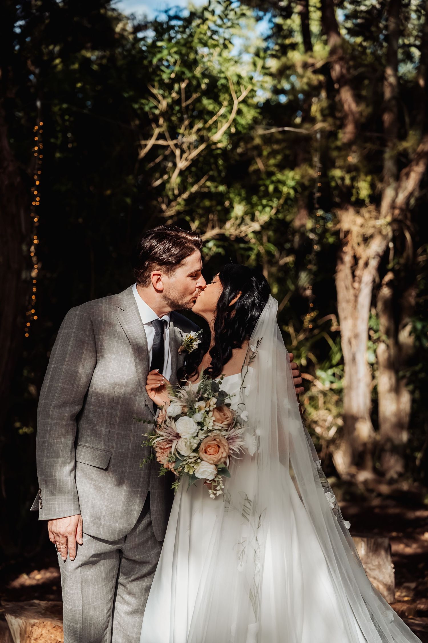 Bride and groom share romantic kiss in dappled sunlight among tall forest trees.