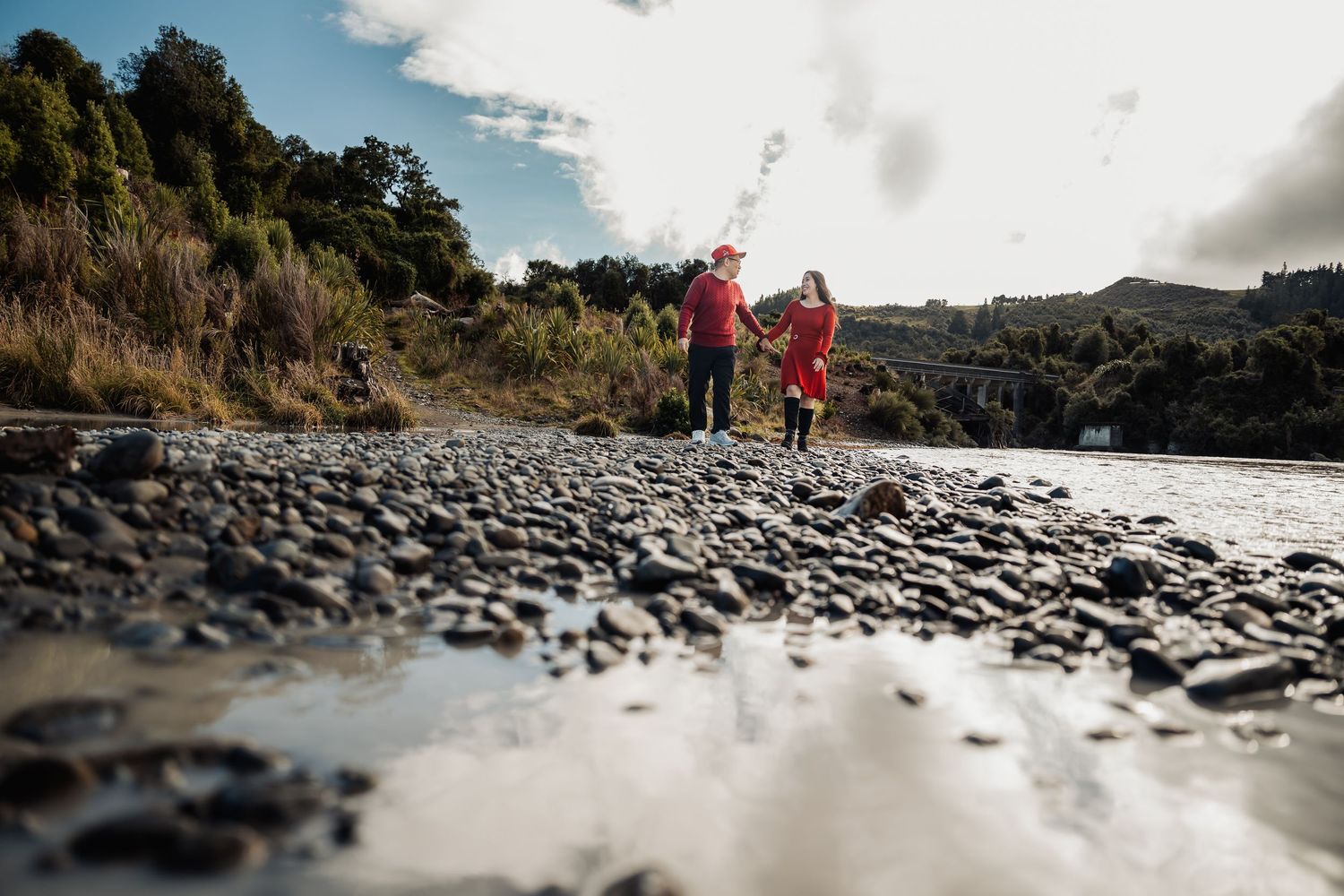 Two people stand on a rocky riverbank surrounded by lush hills and a cloudy sky in New Zealand.